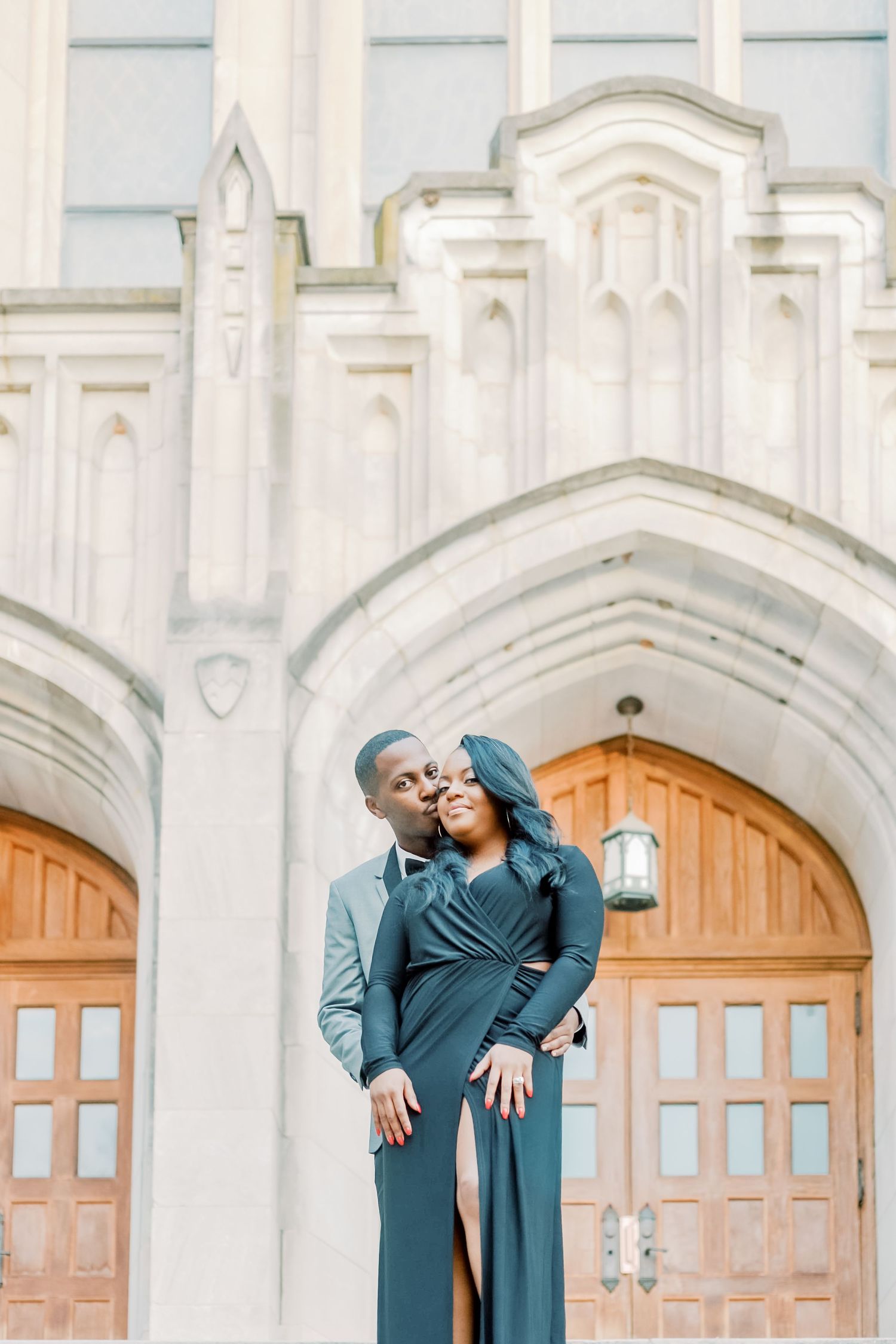 Elegant couple in matching teal outfits embracing before a grand stone archway with wooden doors.