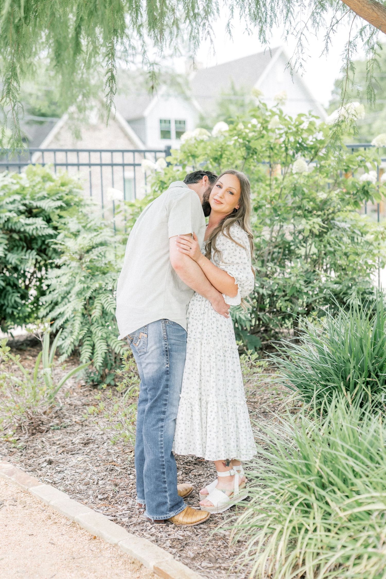 A couple embraces in a lush garden, woman in floral maxi dress smiles while man kisses her cheek.