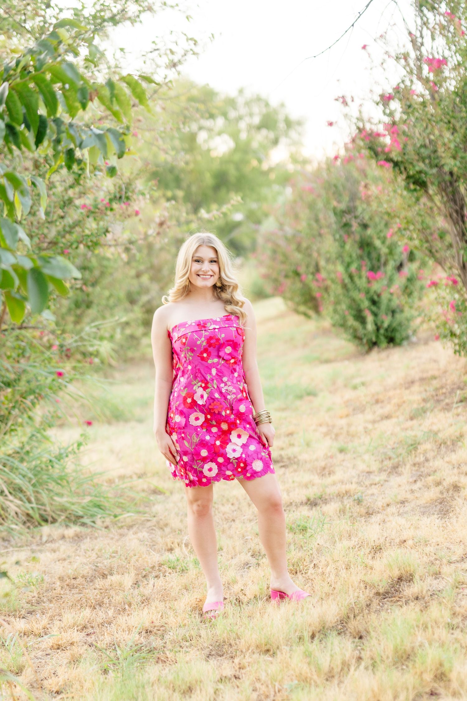 Young blonde woman smiling in pink floral strapless dress standing outdoors among blooming trees.