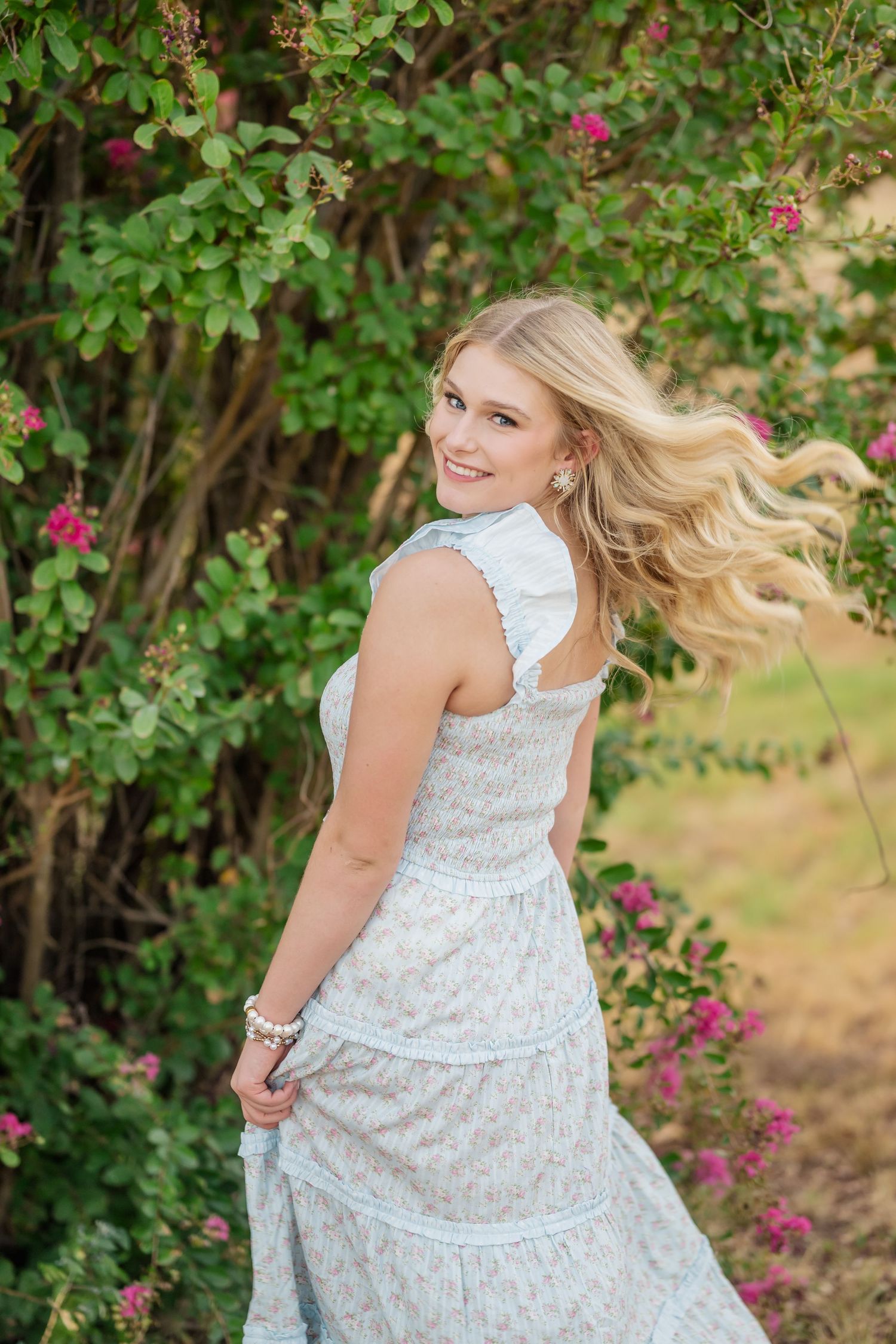 Smiling blonde woman in a floral white dress twirling near blooming pink flowers and lush green bushes outdoors.