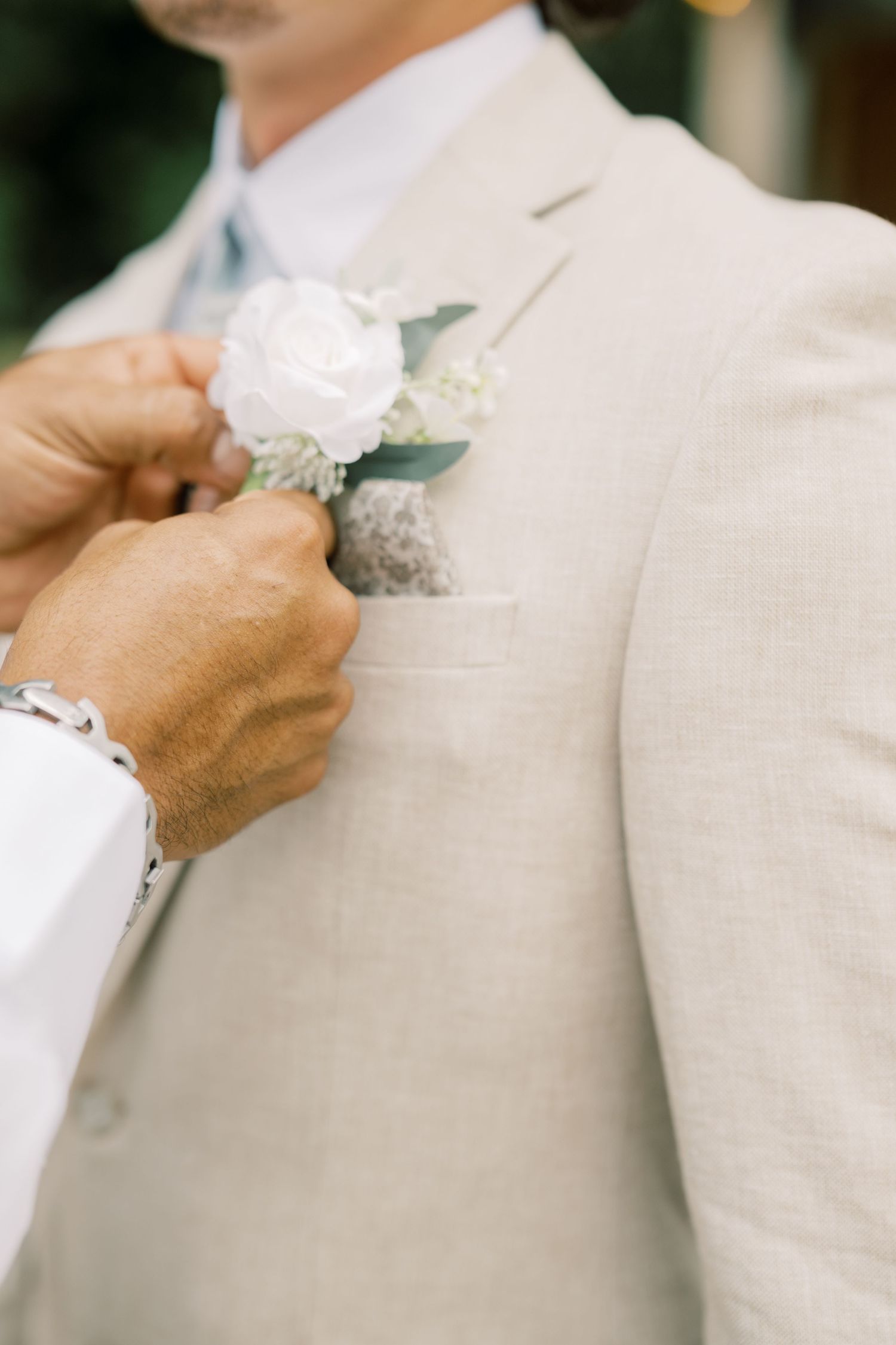 Groom in beige linen suit getting white rose boutonniere pinned to lapel at wedding.