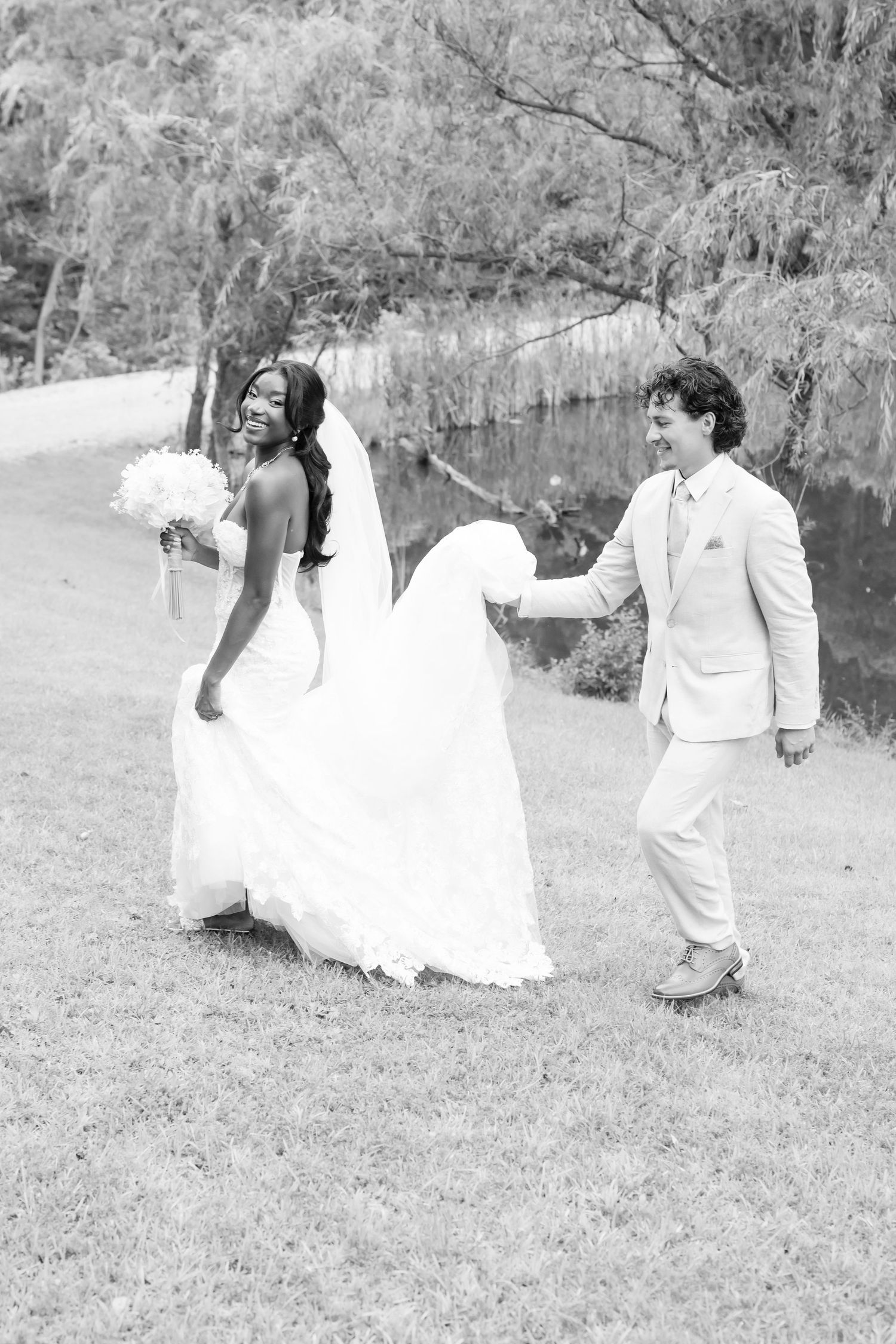 Black and white photo of interracial wedding couple walking by pond, groom holding bride's train as she carries bouquet.