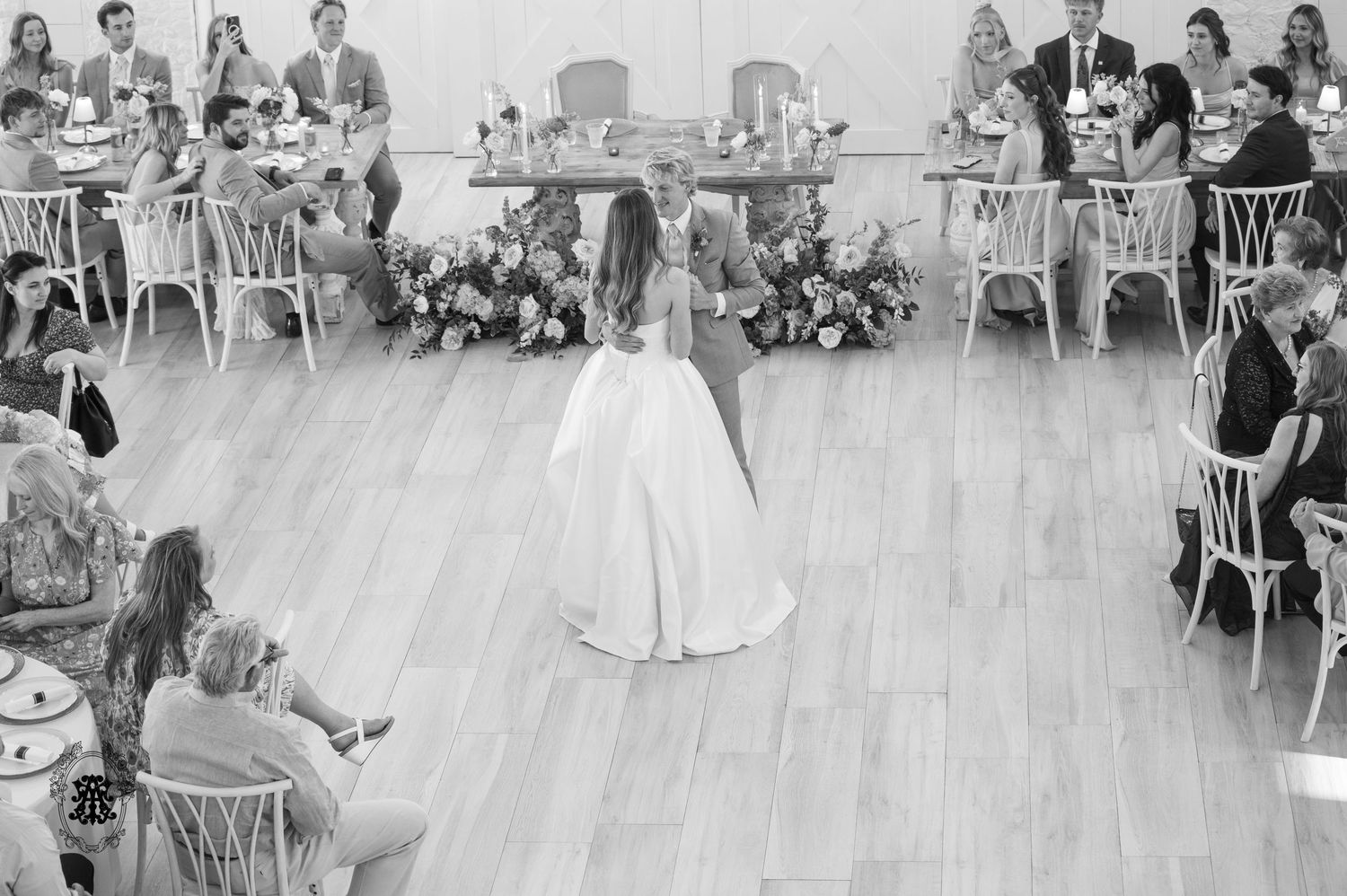 Bride and groom share first dance on wooden floor surrounded by seated wedding guests in elegant reception hall.