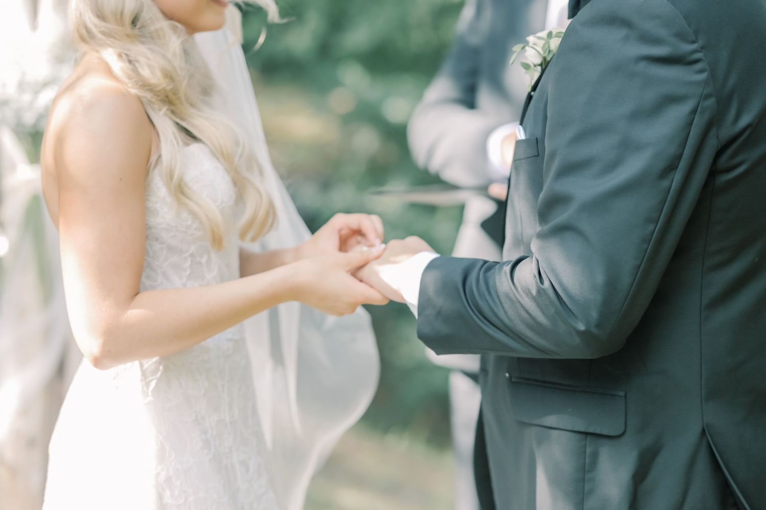 A bride and groom exchange rings during an outdoor wedding ceremony in soft natural light.