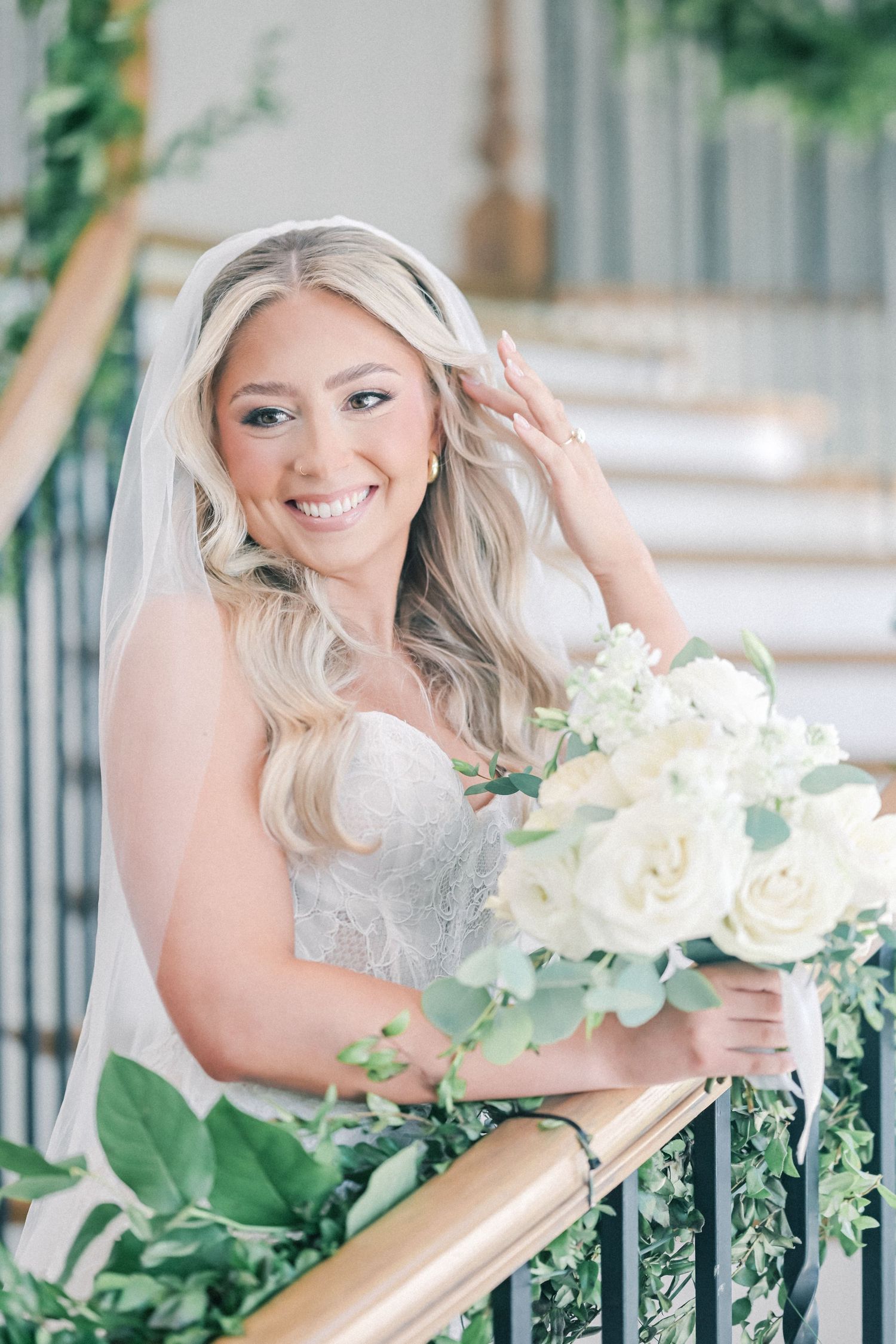 Radiant blonde bride in strapless lace gown holds white rose bouquet while posing on decorated staircase.