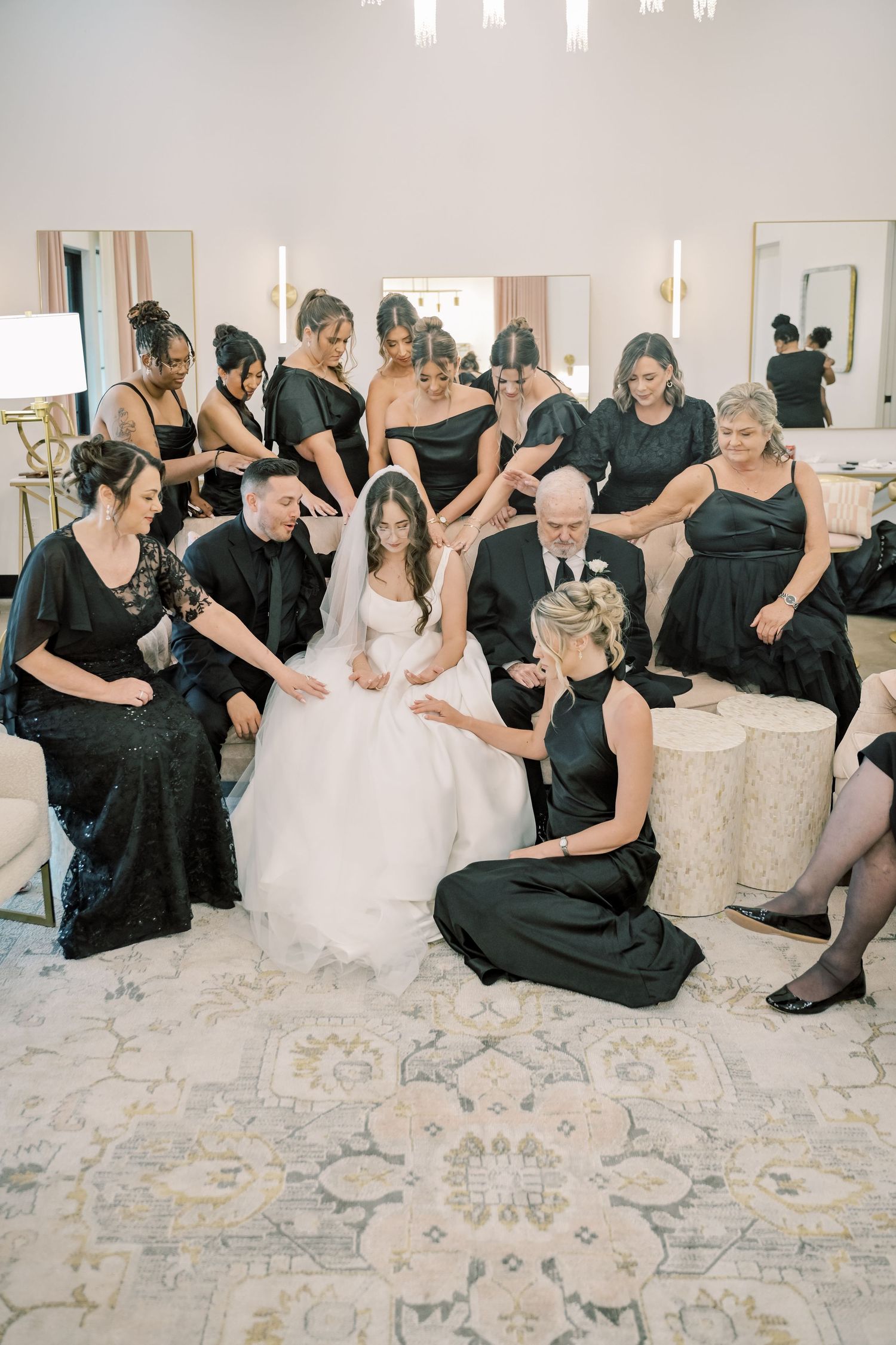 Bride surrounded by bridesmaids in black dresses during an intimate prayer moment in a bright bridal suite.