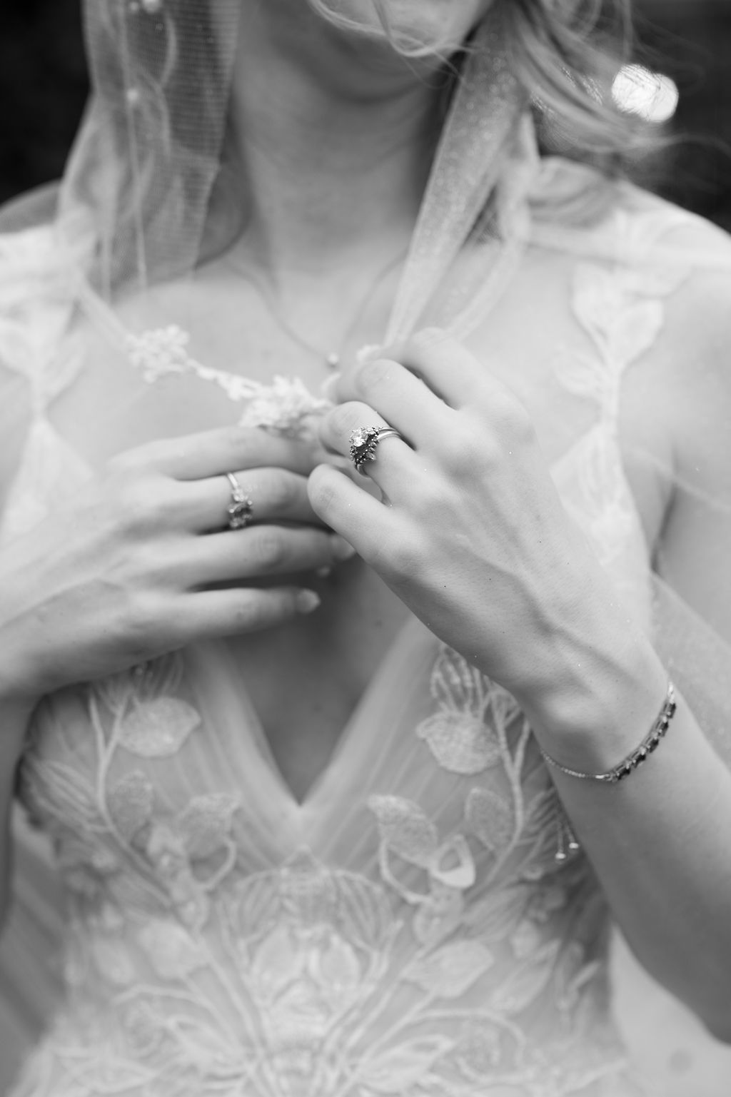 Black and white photo of bride in lace wedding dress adjusting veil, wearing elegant rings and bracelet.