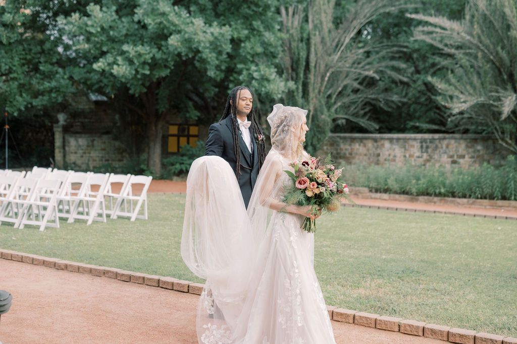 Bride and groom walking outdoors at elegant garden wedding venue with white chairs and lush greenery.
