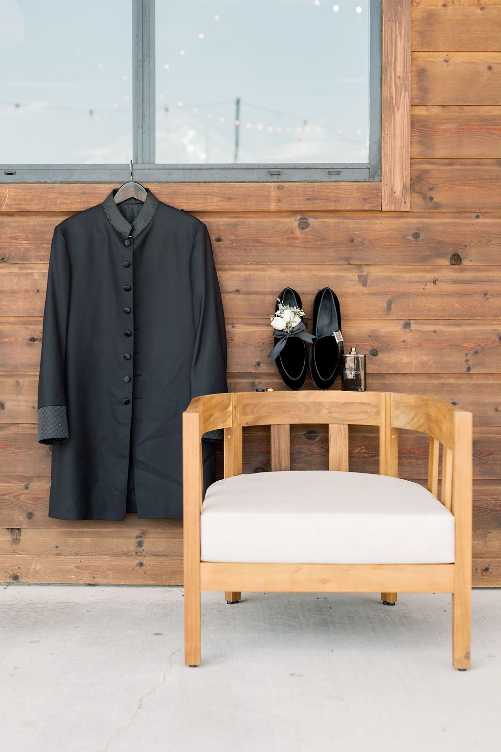 Groom wedding attire flat lay featuring black long coat on hanger, dress shoes, and boutonniere on wooden chair.
