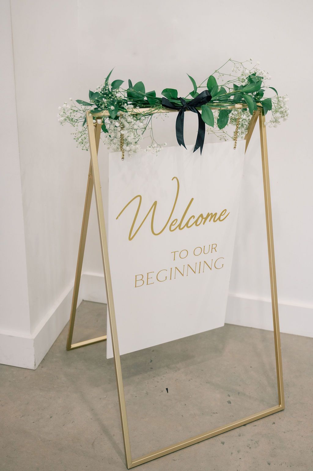 Gold-framed wedding welcome sign reading Welcome to Our Beginning adorned with greenery and white flowers.