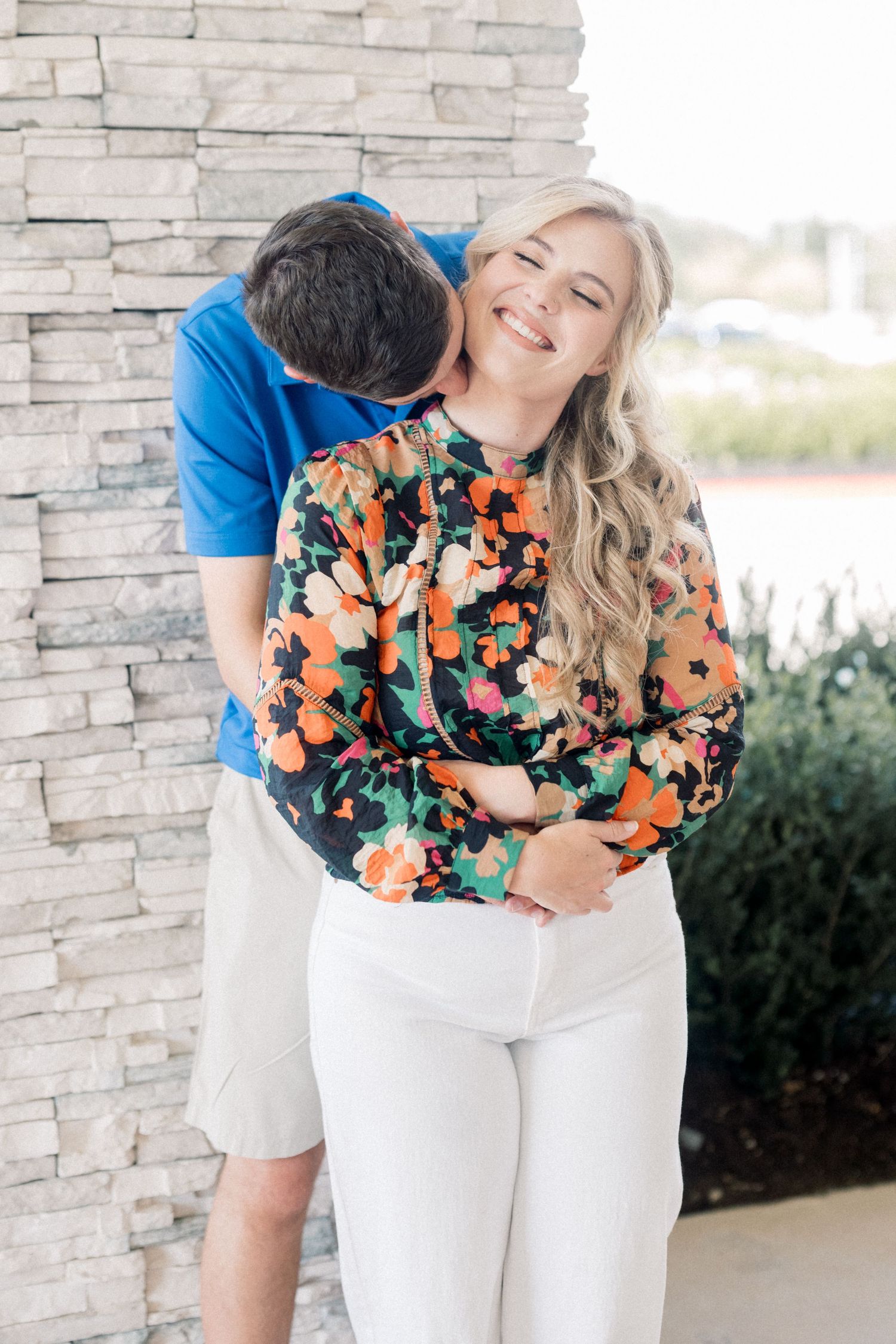 Man kissing smiling woman on cheek against stone wall, she wears a vibrant floral top and white pants.