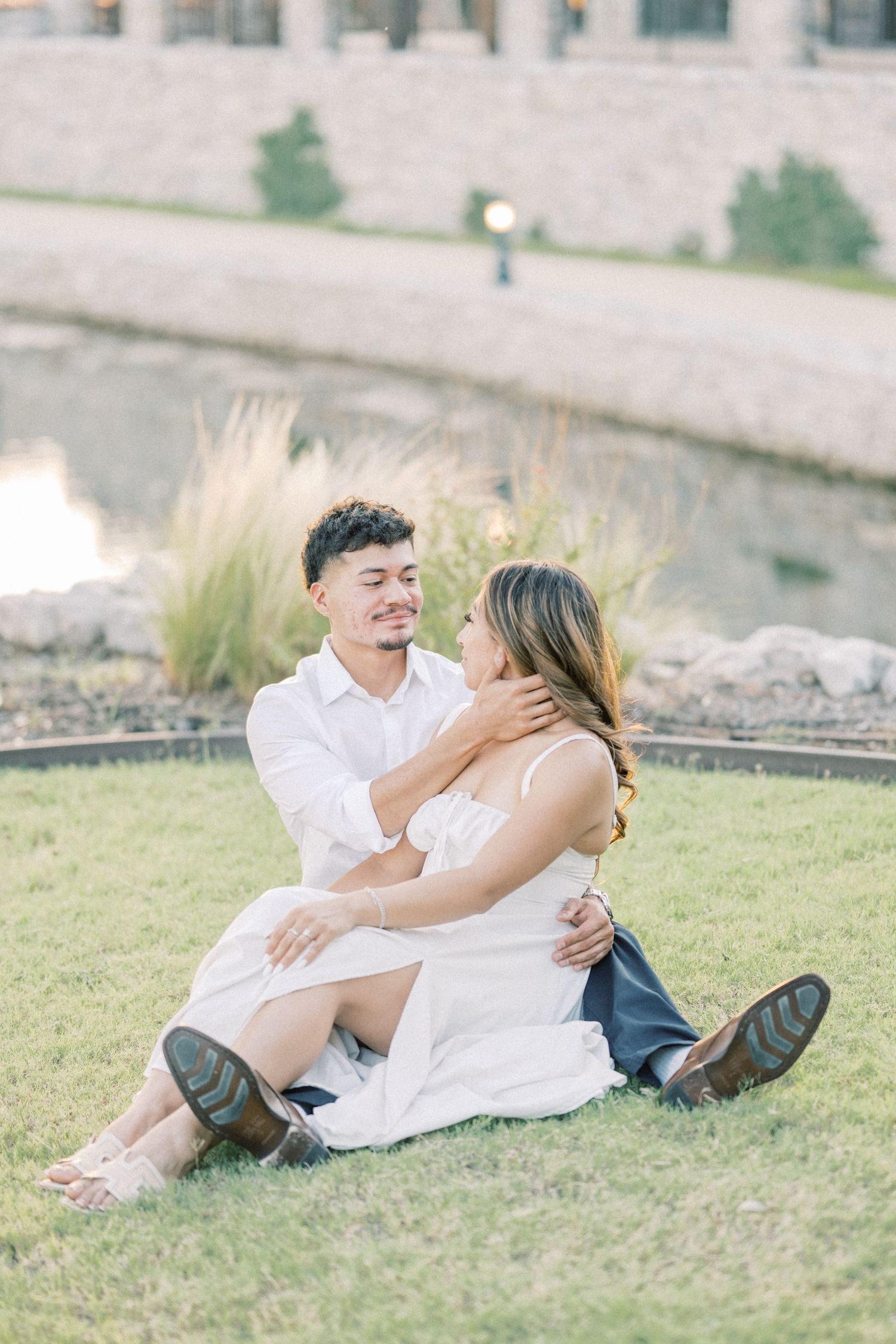 A couple sits together on green grass, gazing at each other tenderly during a golden hour photo session.