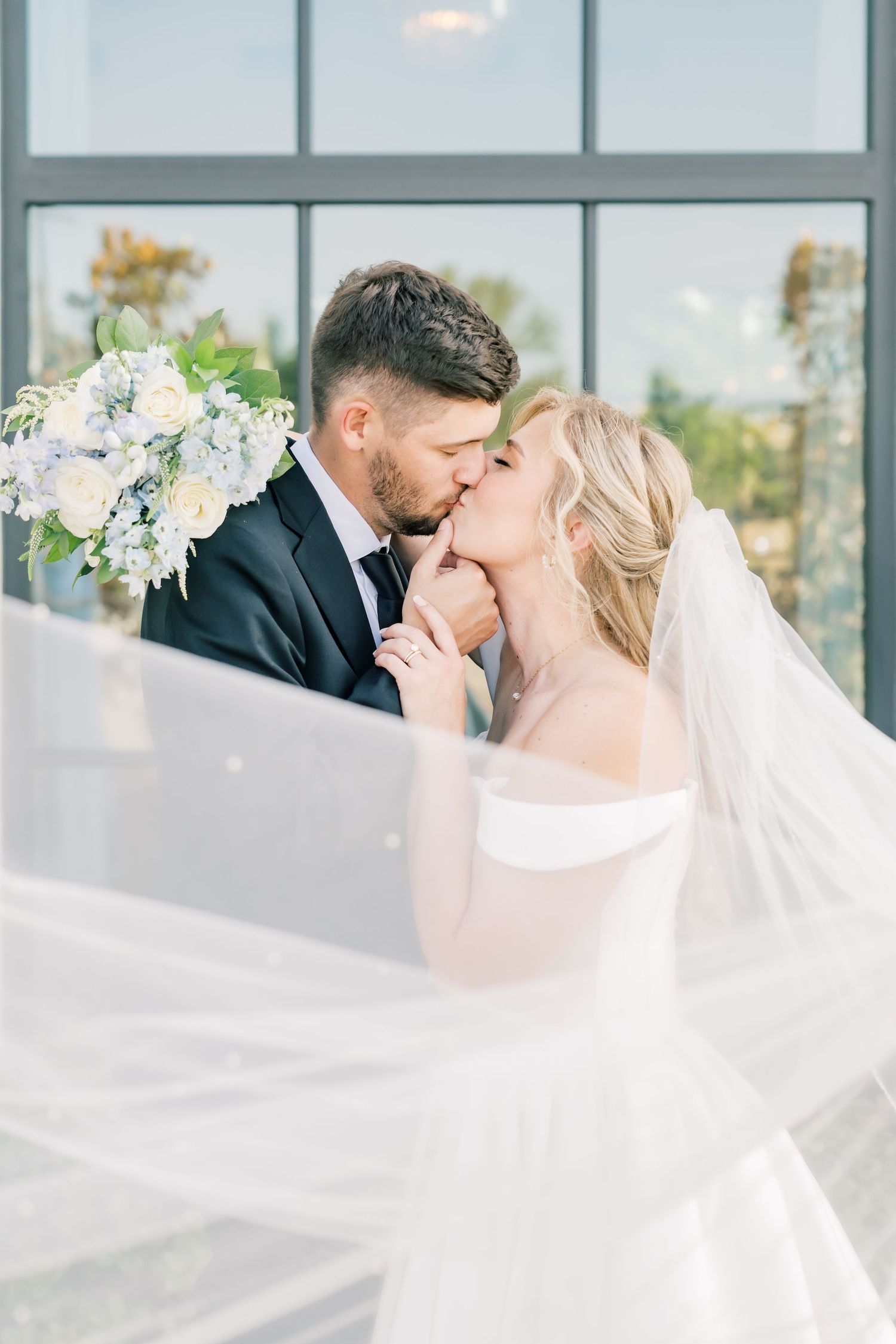 Bride and groom share romantic kiss with flowing veil and white floral bouquet by large windows.