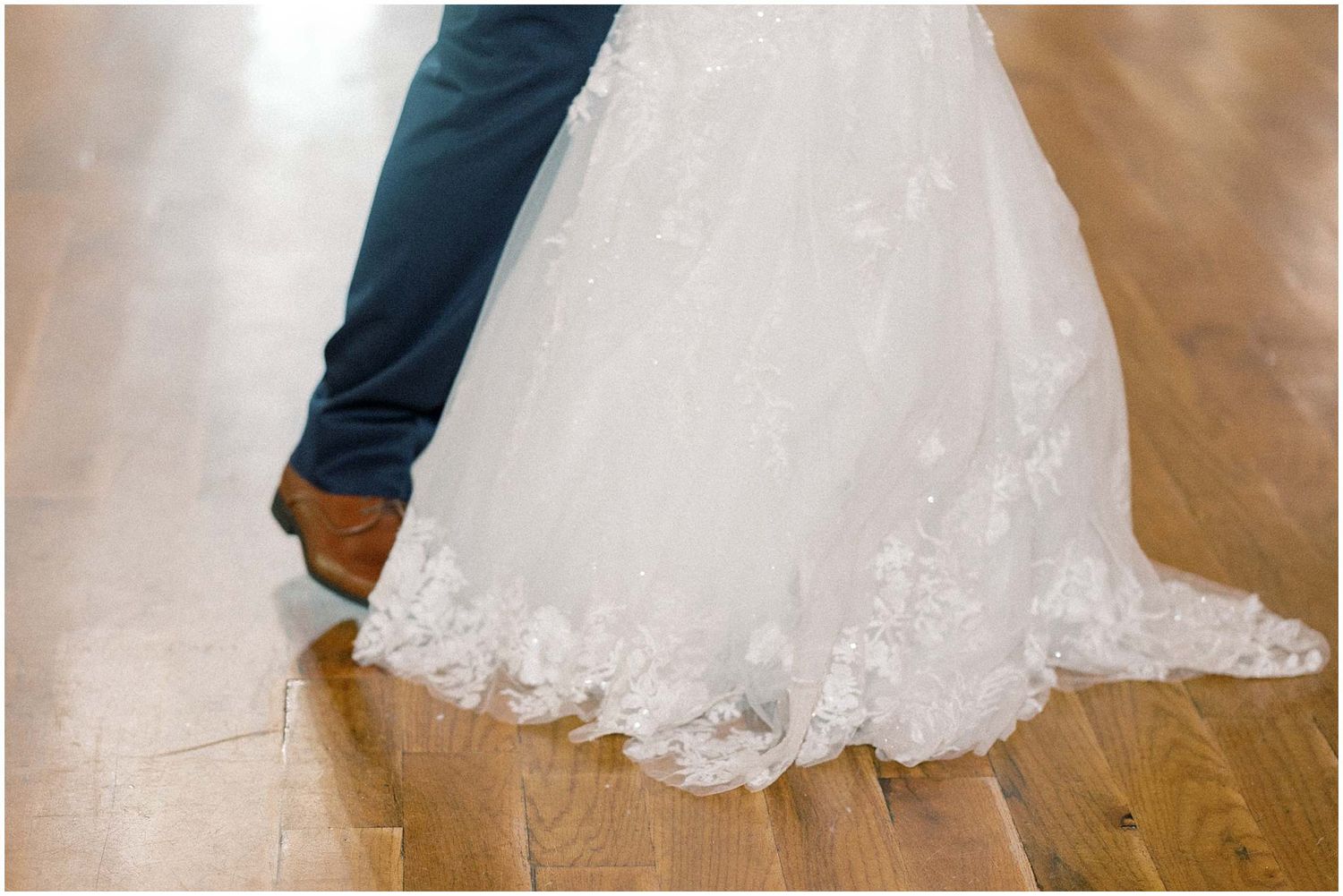 A close-up view of a white lace wedding dress hem and skirt flowing elegantly across a wooden floor.