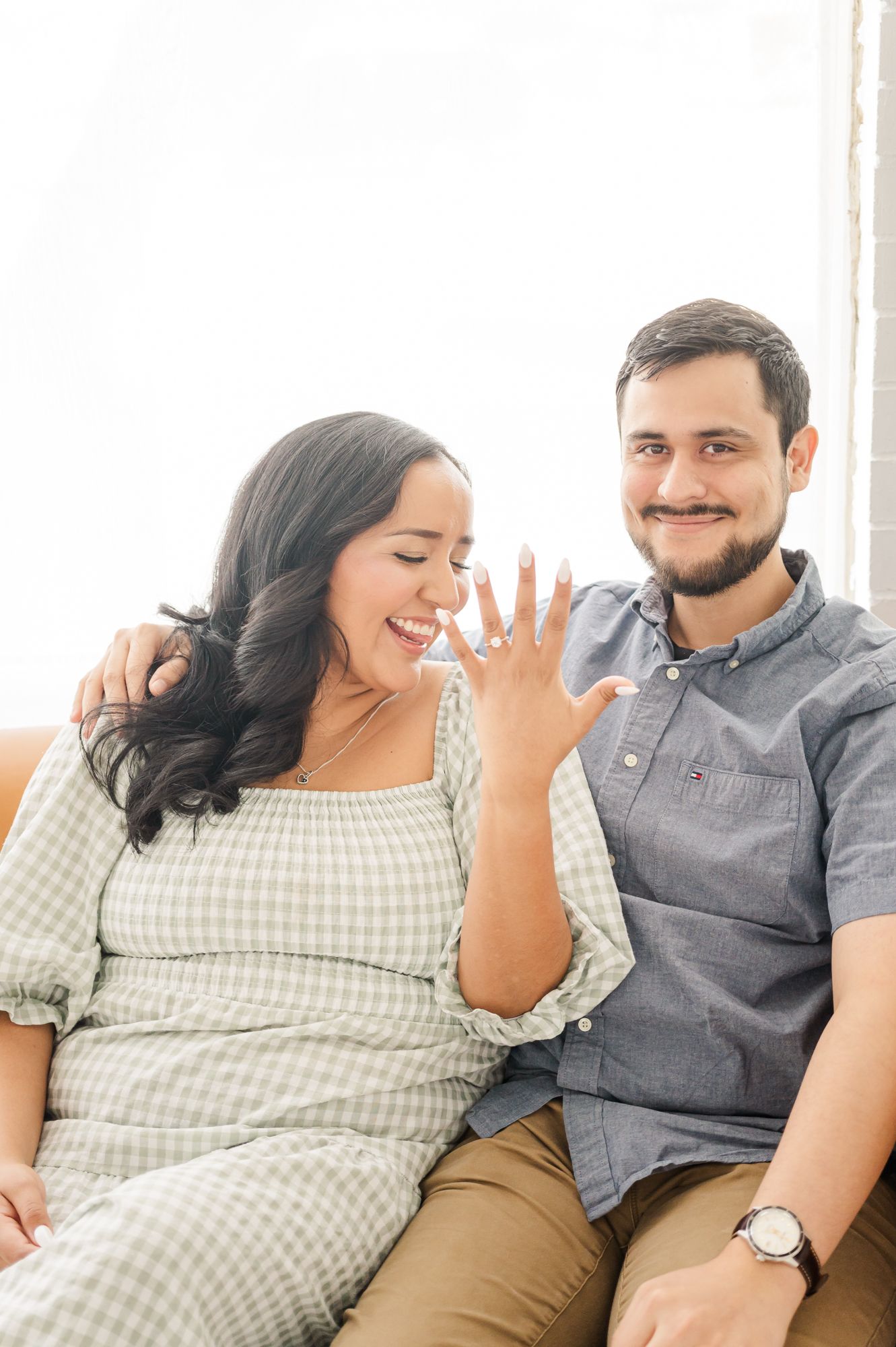 A couple sits together on a sofa while showing off an engagement ring.