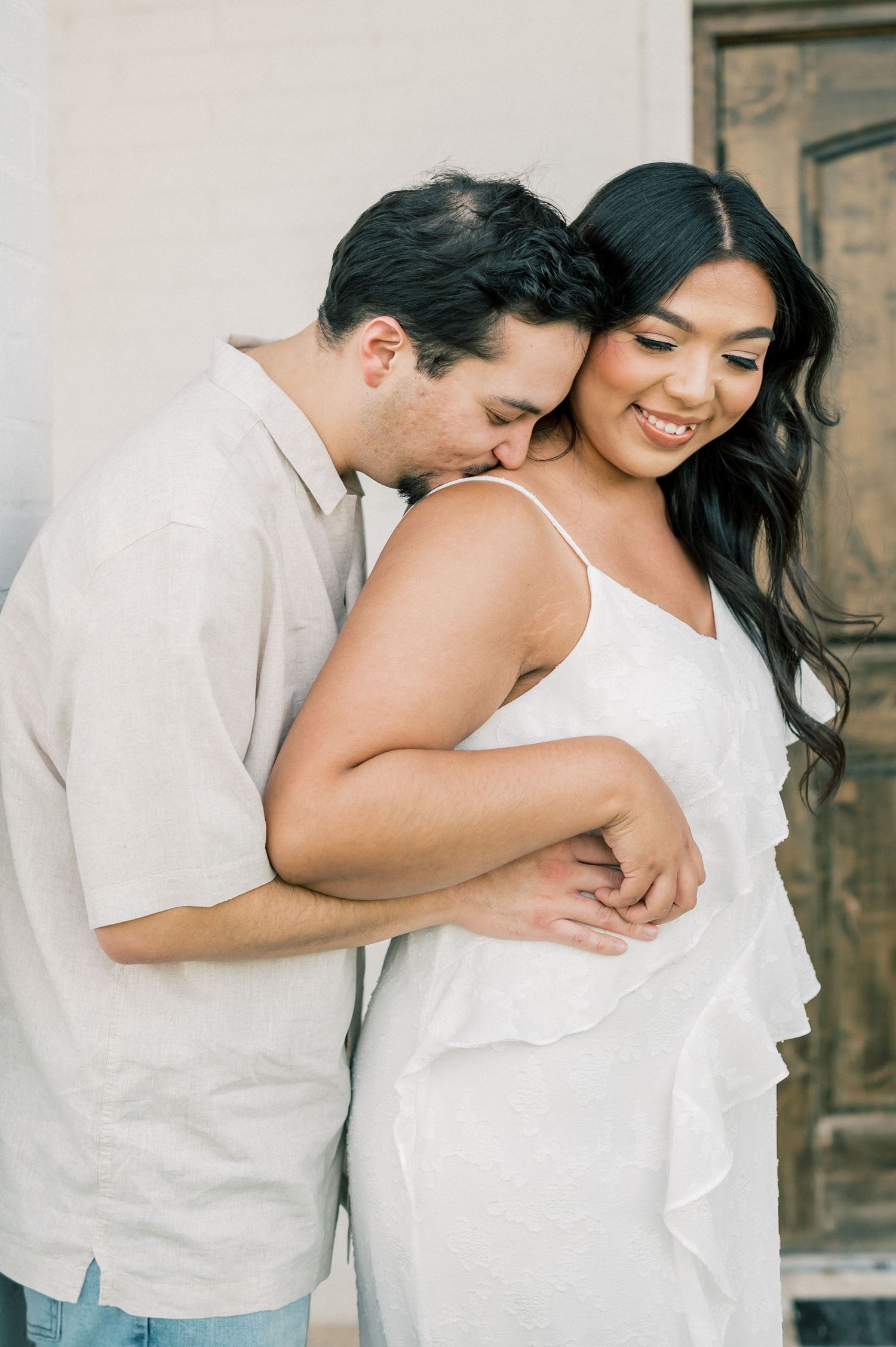A romantic couple in white outfits sharing an intimate moment during a maternity photoshoot against a rustic backdrop.
