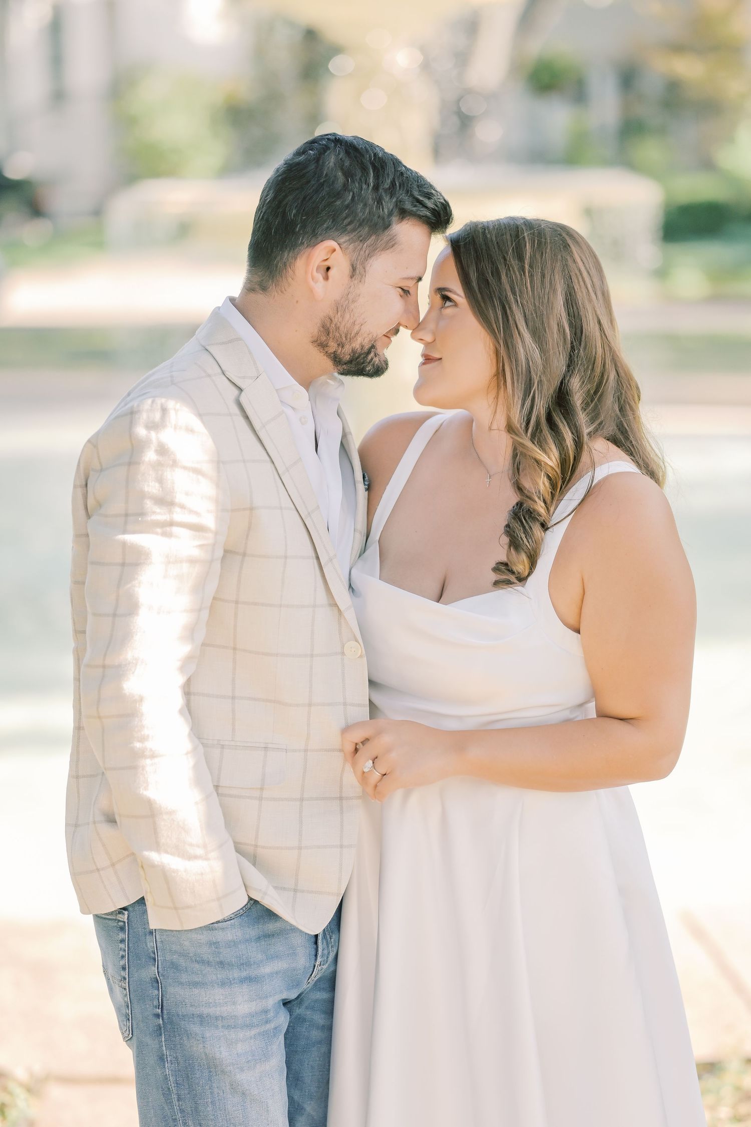 Couple touching noses in a tender moment, the man in a plaid blazer and the woman in a white sundress, during an engagement shoot.