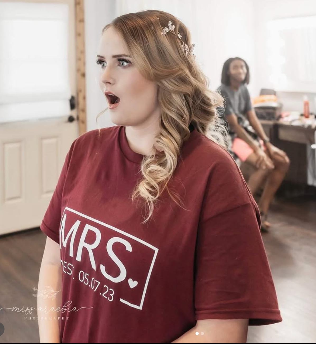 Woman in maroon MRS. shirt dated 05.17.23 with floral hair accessory, looking surprised in bright room.