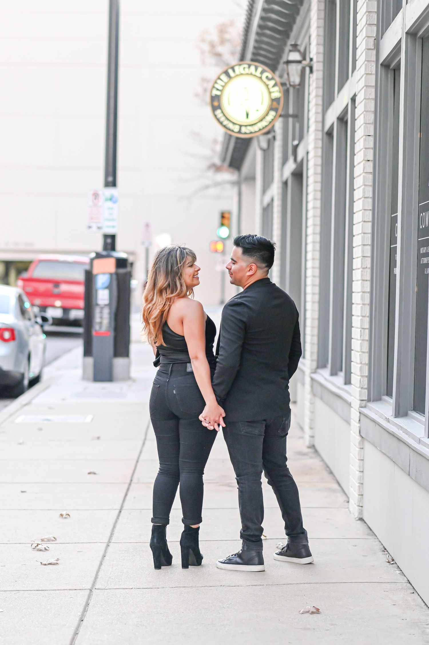 Couple in matching black outfits holding hands while looking at each other on an urban downtown sidewalk.