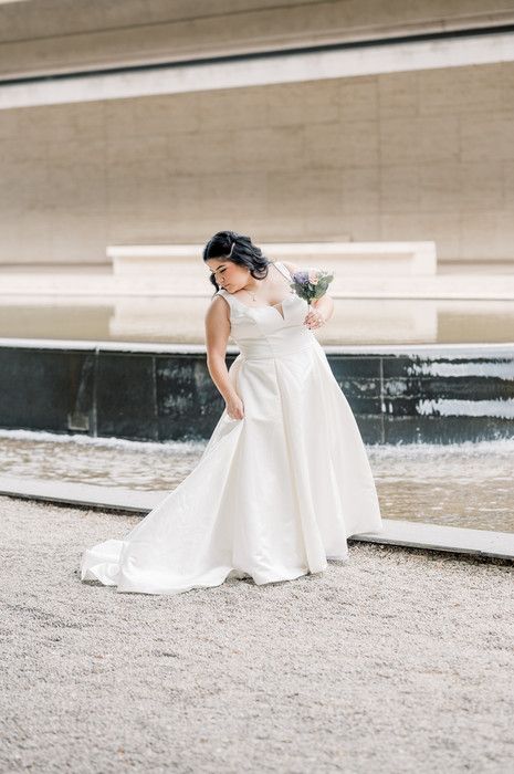 Bride in elegant white ball gown holding bouquet stands near fountain on gravel path at outdoor wedding venue.