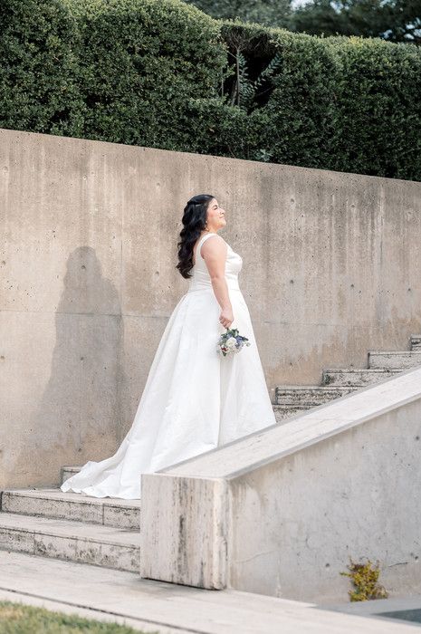 Bride in white ball gown holding bouquet stands on stone steps with lush green hedges backdrop.