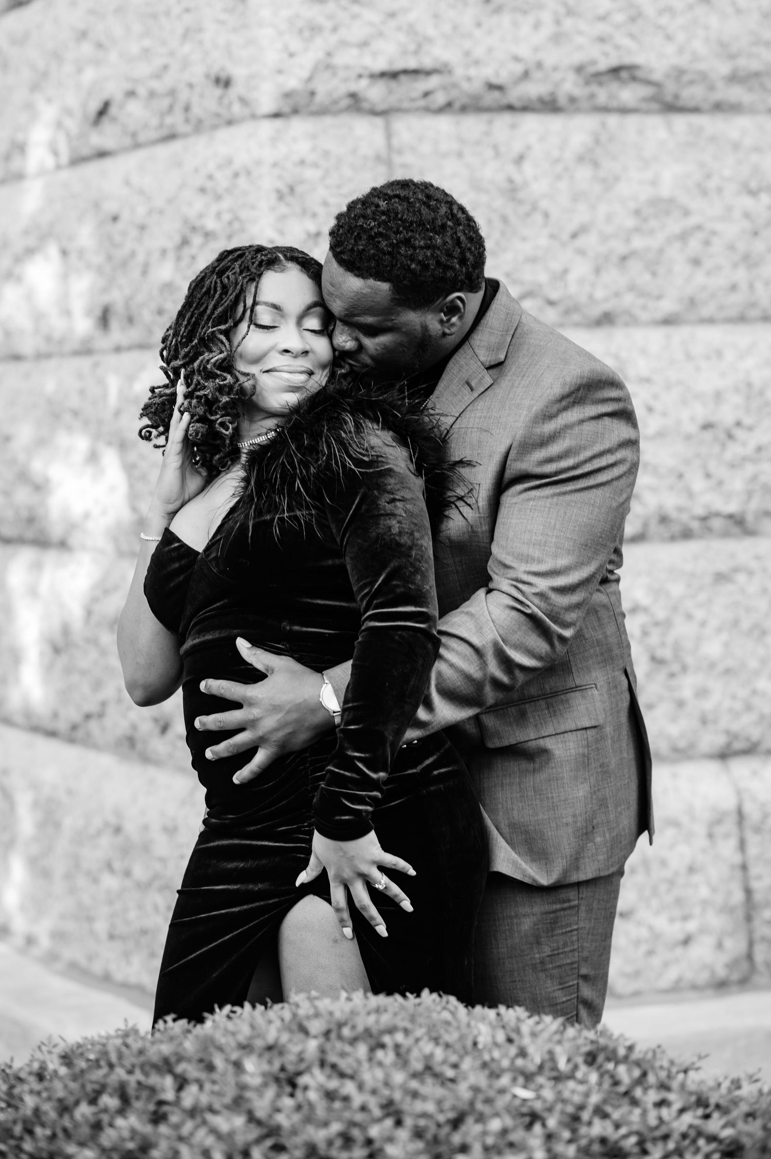 Black and white photo of a couple embracing, man kissing woman's cheek, both dressed elegantly outdoors.