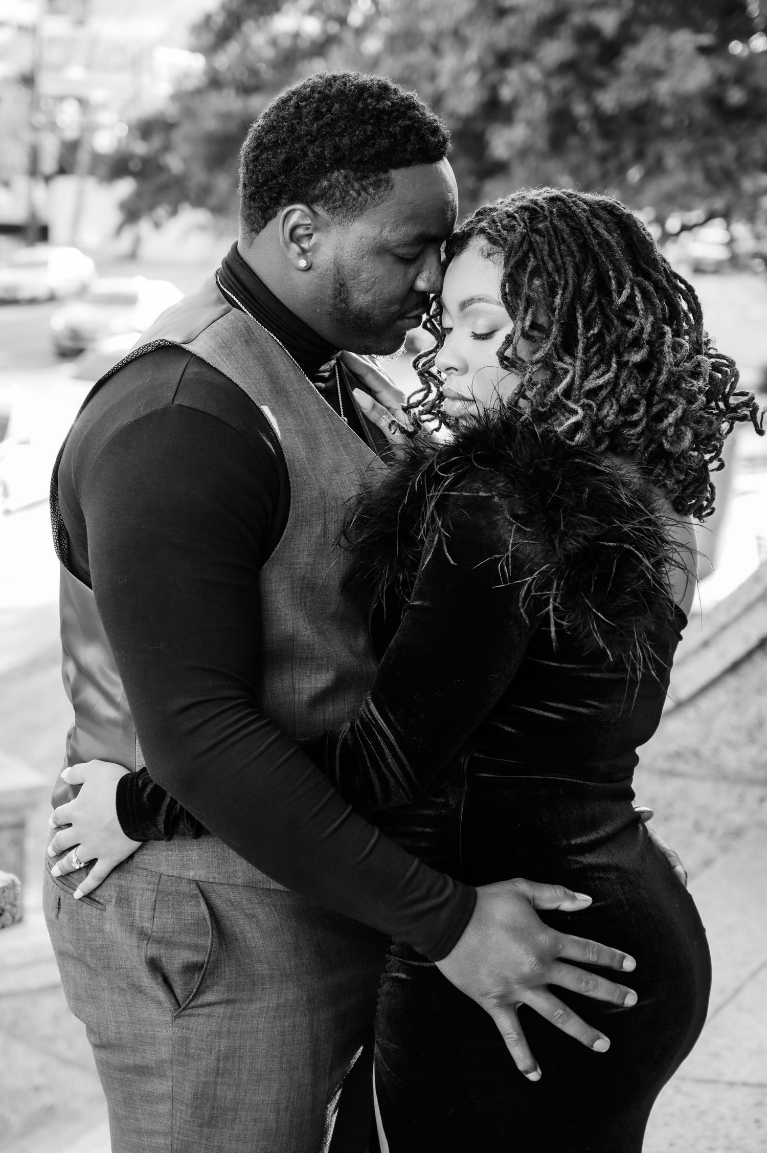 Black and white photo of a couple embracing and kissing outdoors, man in vest and woman in black velvet dress.