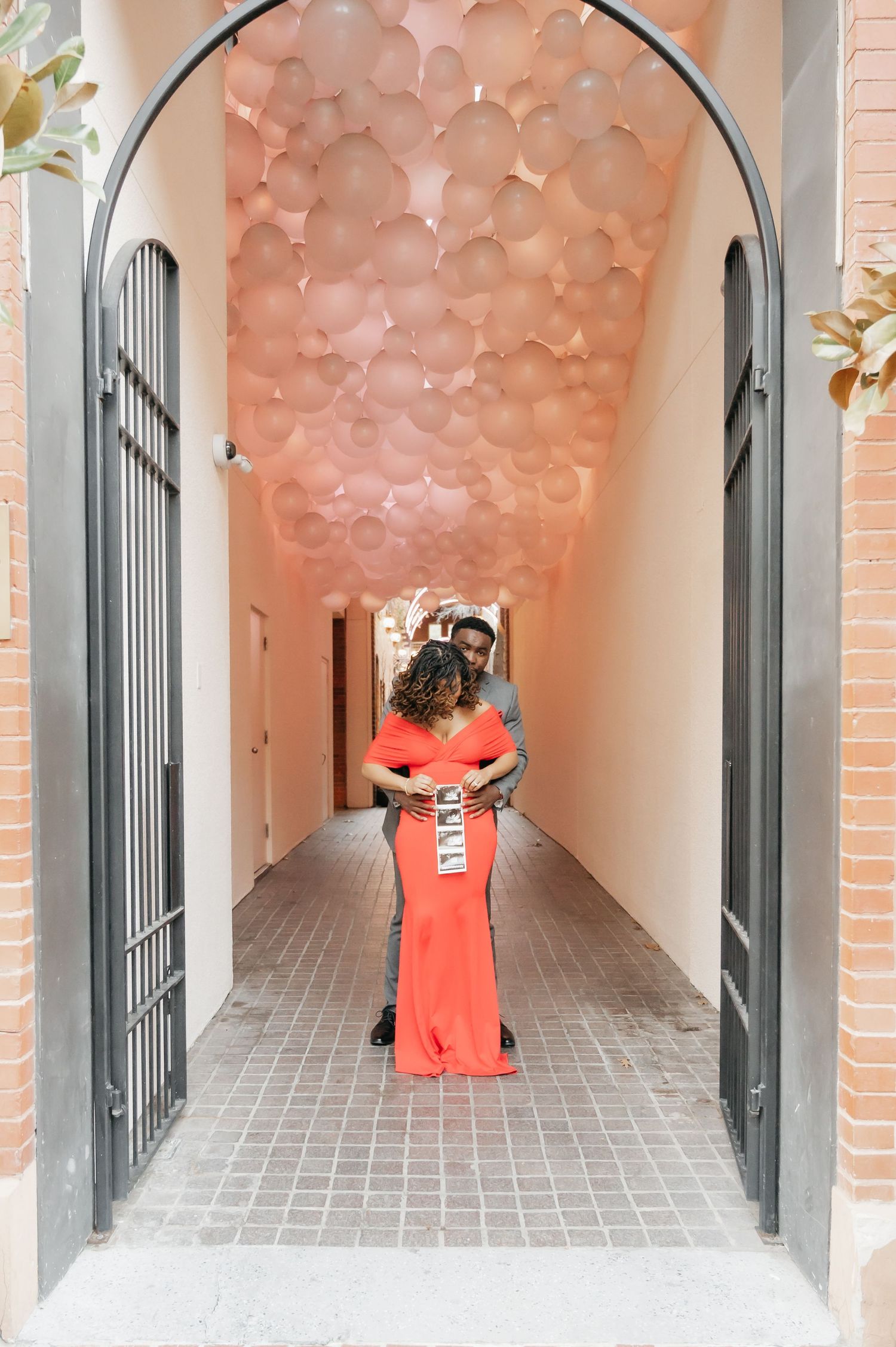 Couple embracing in a romantic pink balloon-lined hallway, woman in red gown.