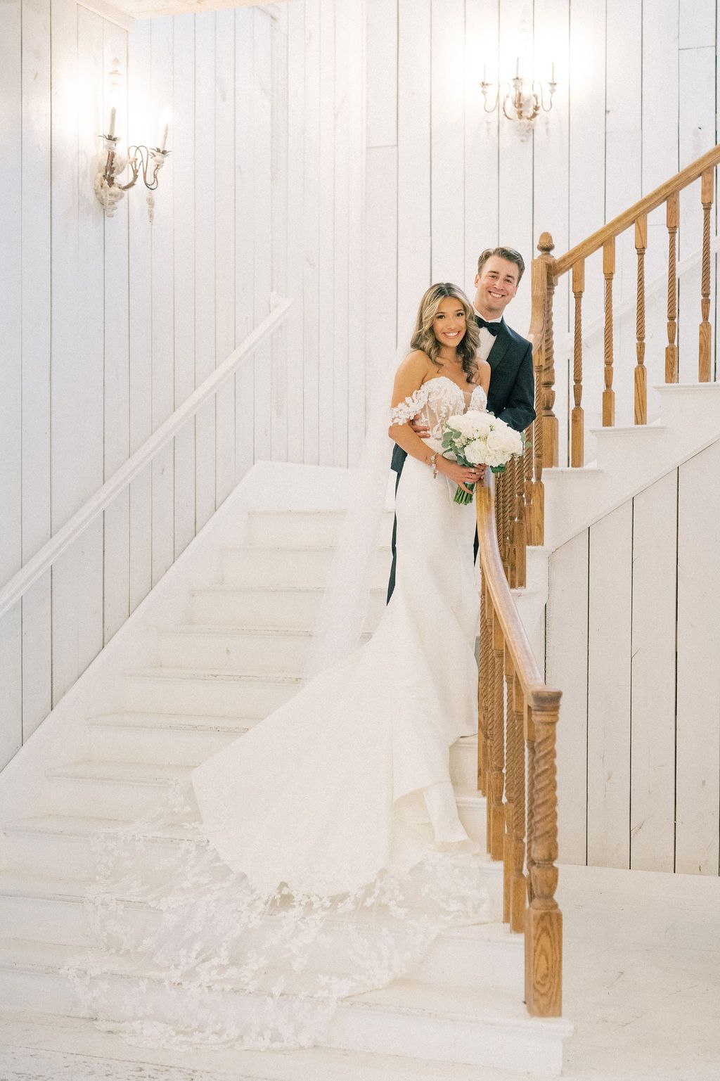Smiling bride and groom pose on white staircase, her off-shoulder gown and lace train cascading down the steps.