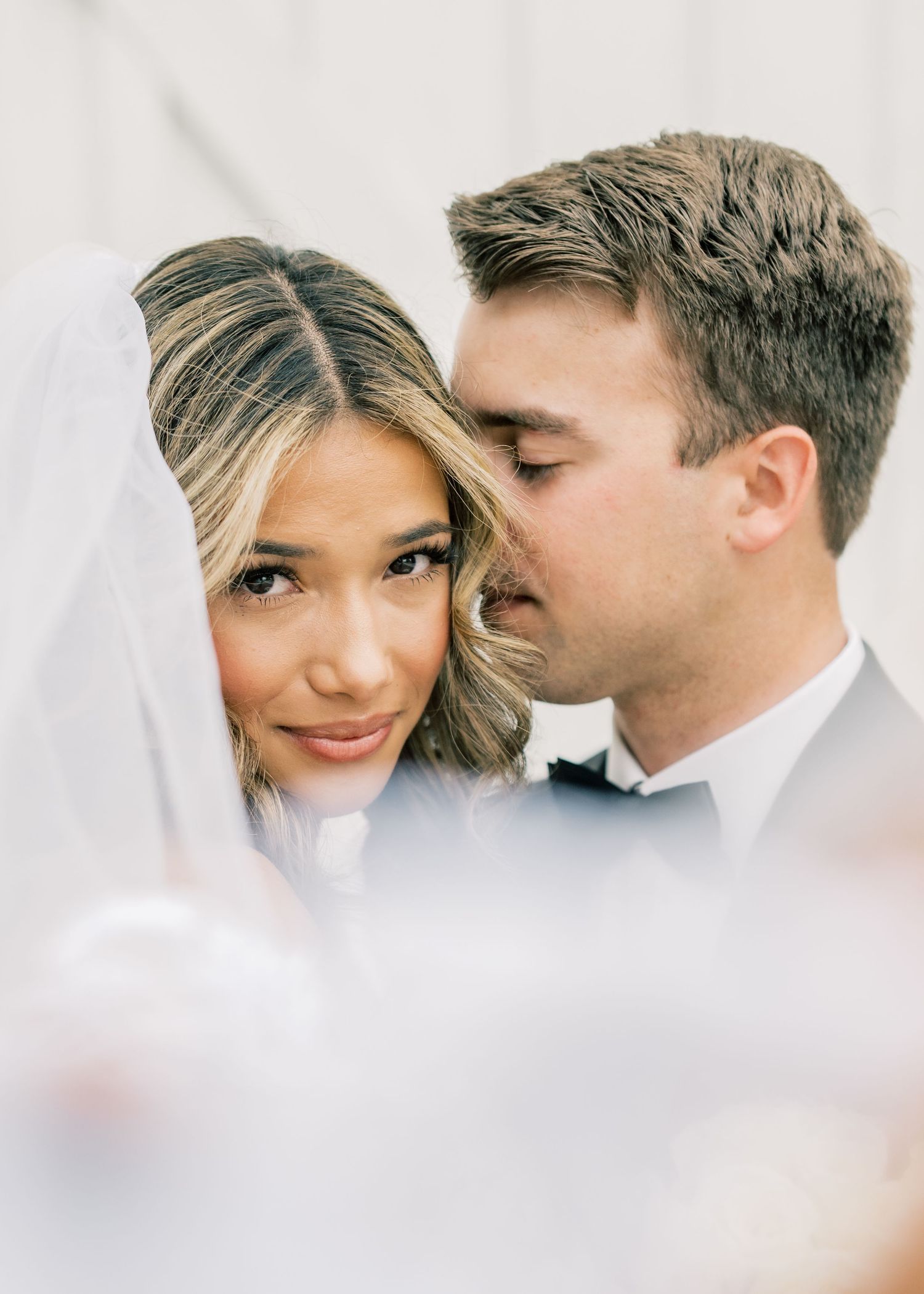 Groom tenderly kisses bride on the cheek as she smiles under her flowing white wedding veil.