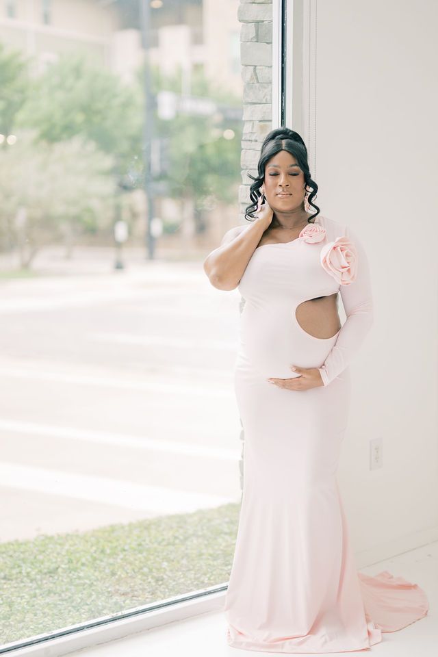 Pregnant woman in pink one-shoulder cutout gown with floral detail by bright window.