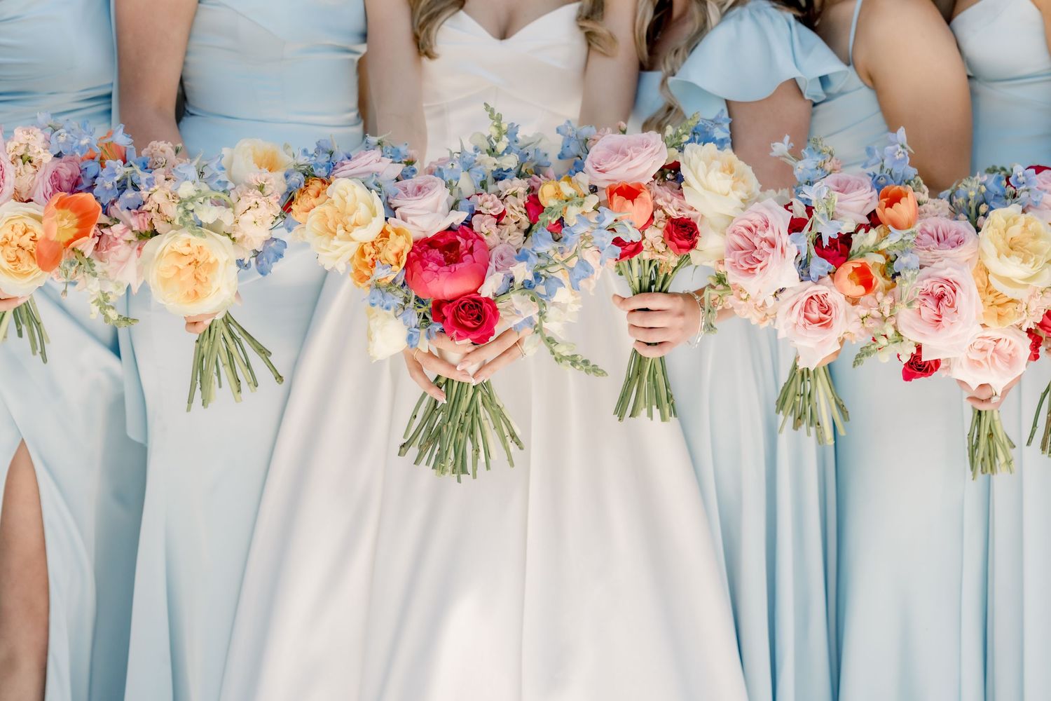 Bridesmaids in light blue gowns holding colorful spring bouquets with pink, peach, and red flowers beside a bride.