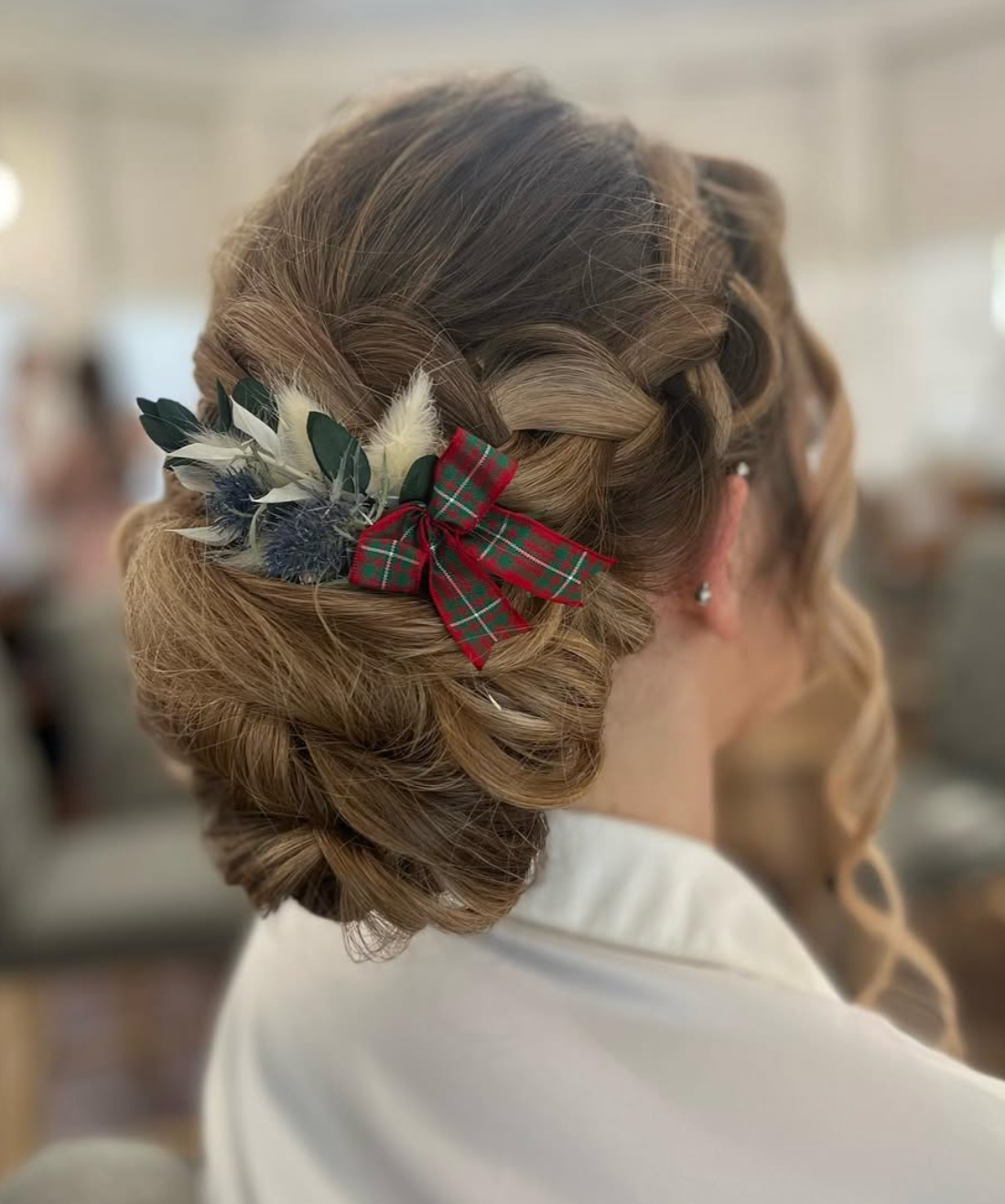 Elegant bridal updo with braided bun, tartan bow, and dried floral accents on medium brown hair.