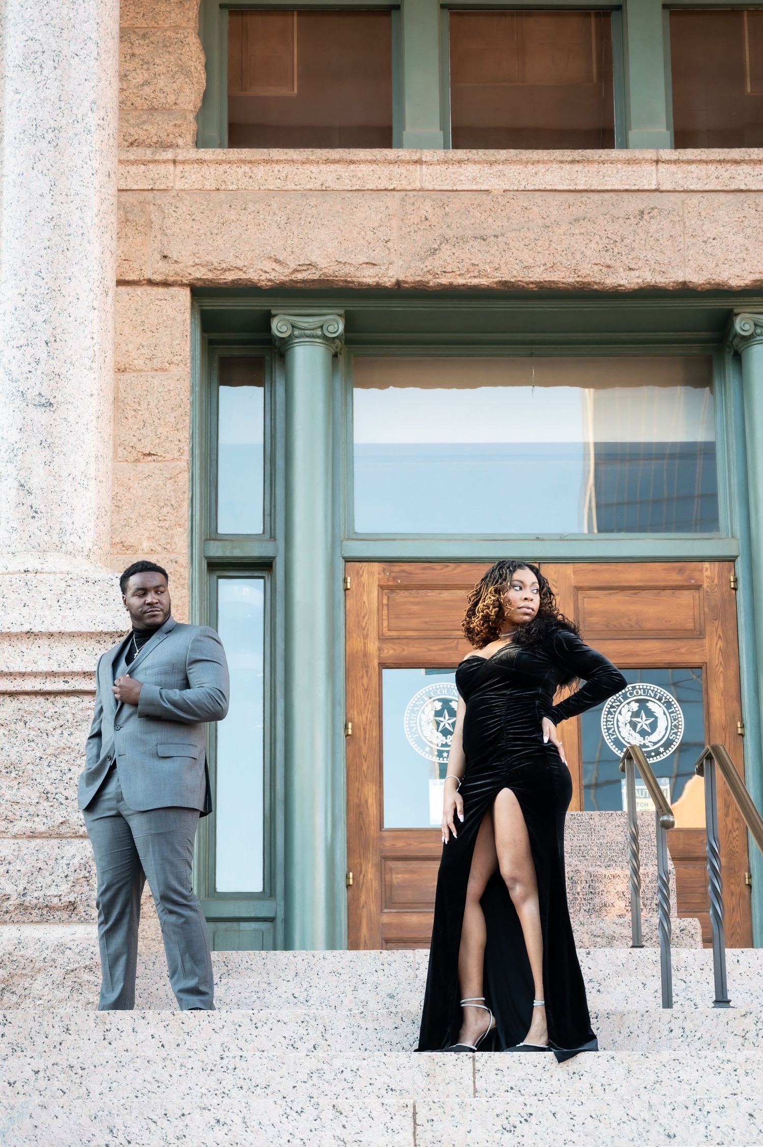 Elegant couple posing on steps, man in gray suit and woman in black velvet gown with slit, before a classic building.
