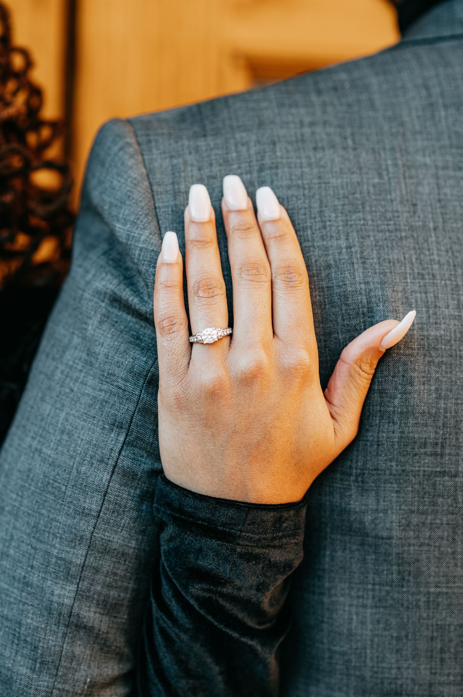 Close-up of a hand with white manicured nails wearing a diamond engagement ring resting on a gray suit jacket.