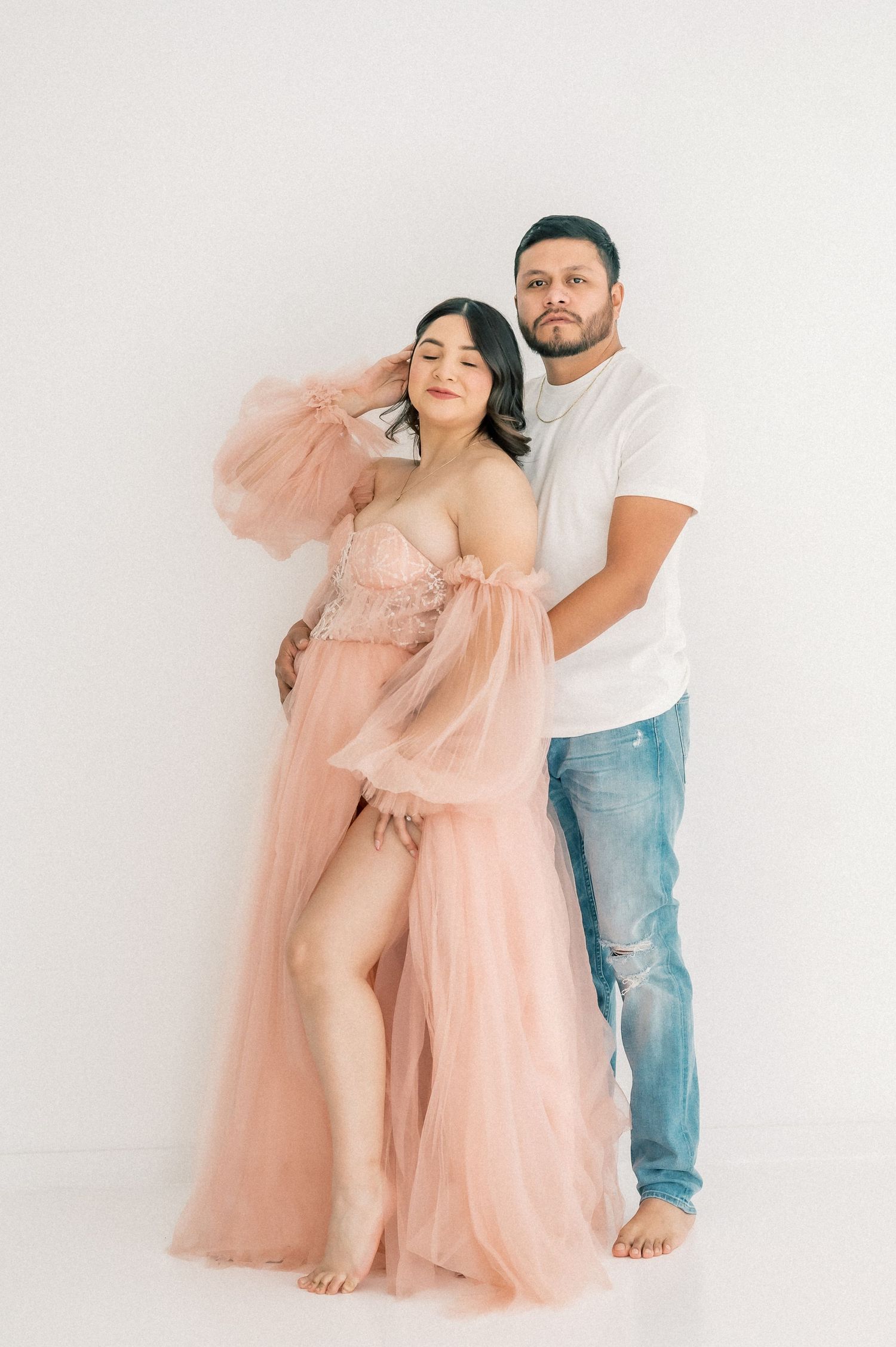 Expecting couple in a studio, woman in blush tulle gown and man in white tee and jeans, maternity portrait.