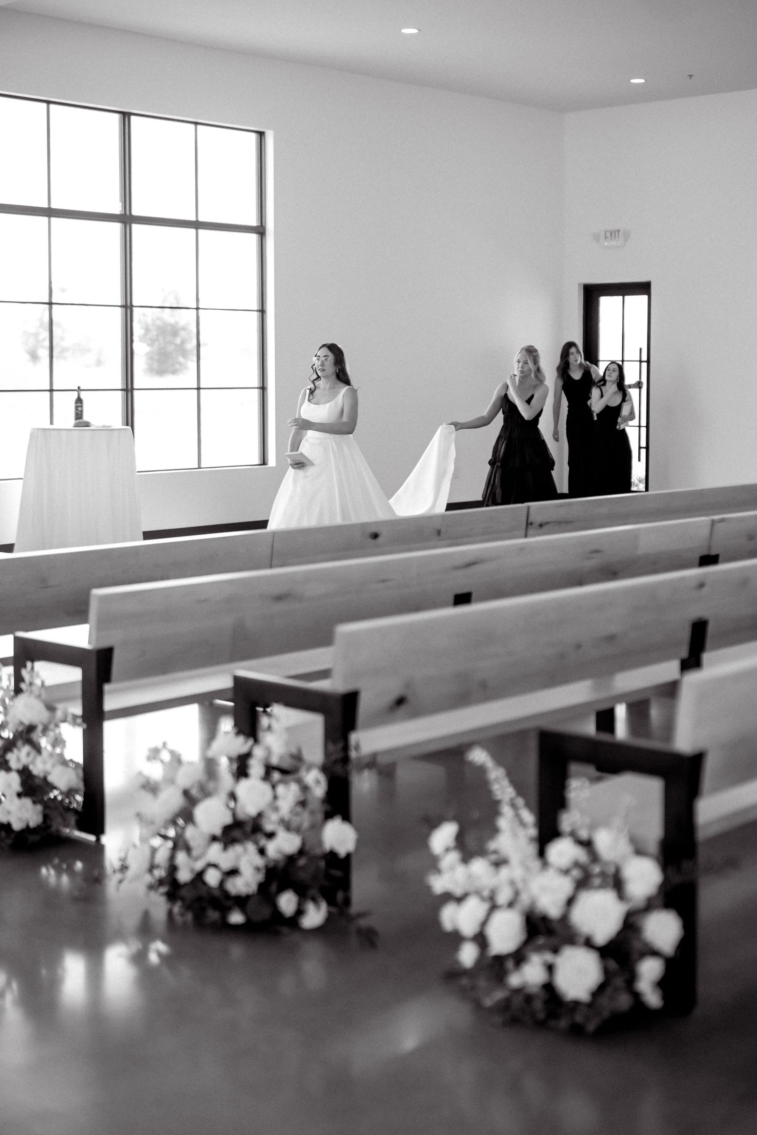Black and white photo of bride walking down chapel aisle with bridesmaids carrying her train.