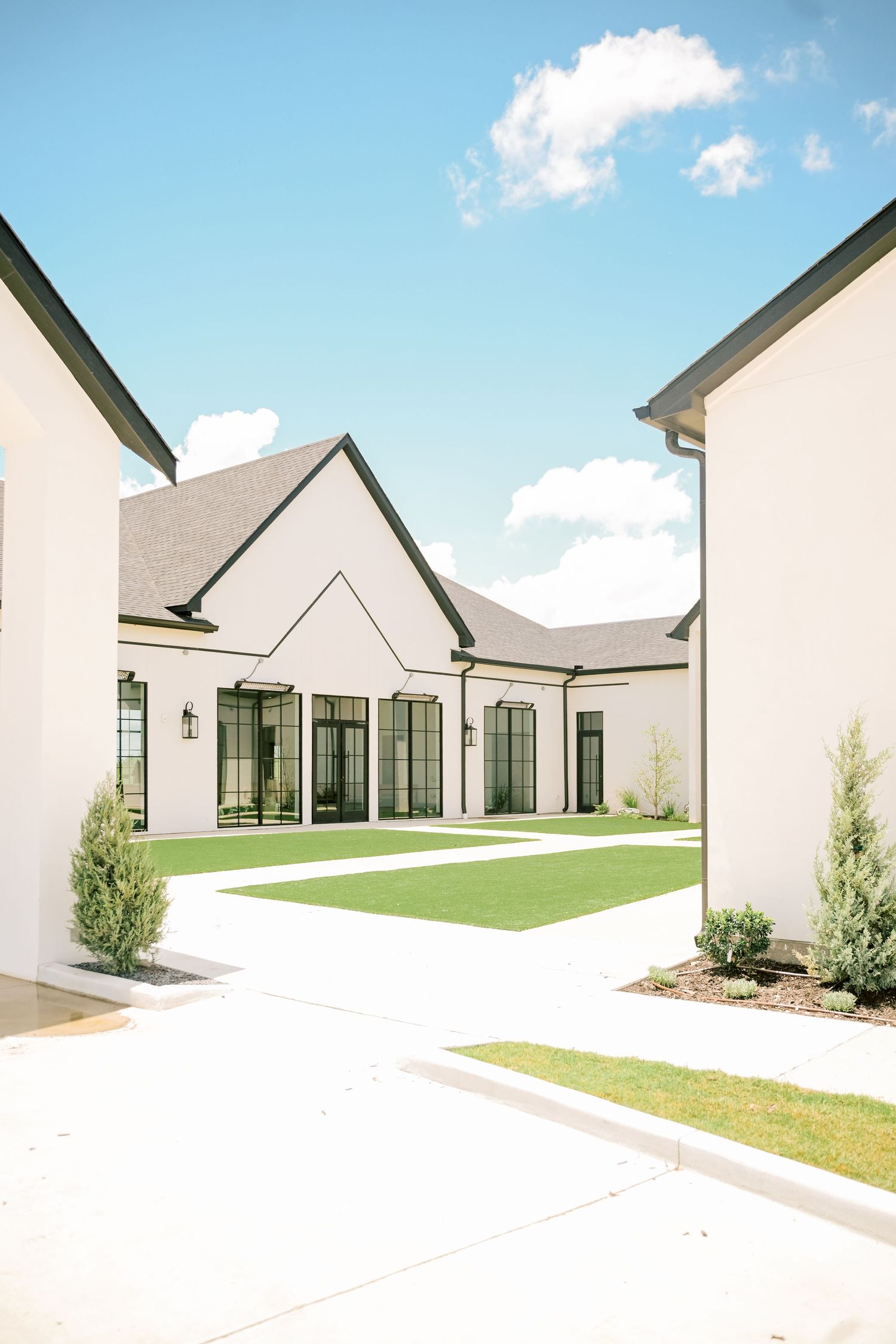 Modern white wedding venue exterior with black-trimmed windows, manicured lawn, and elegant courtyard under blue sky.