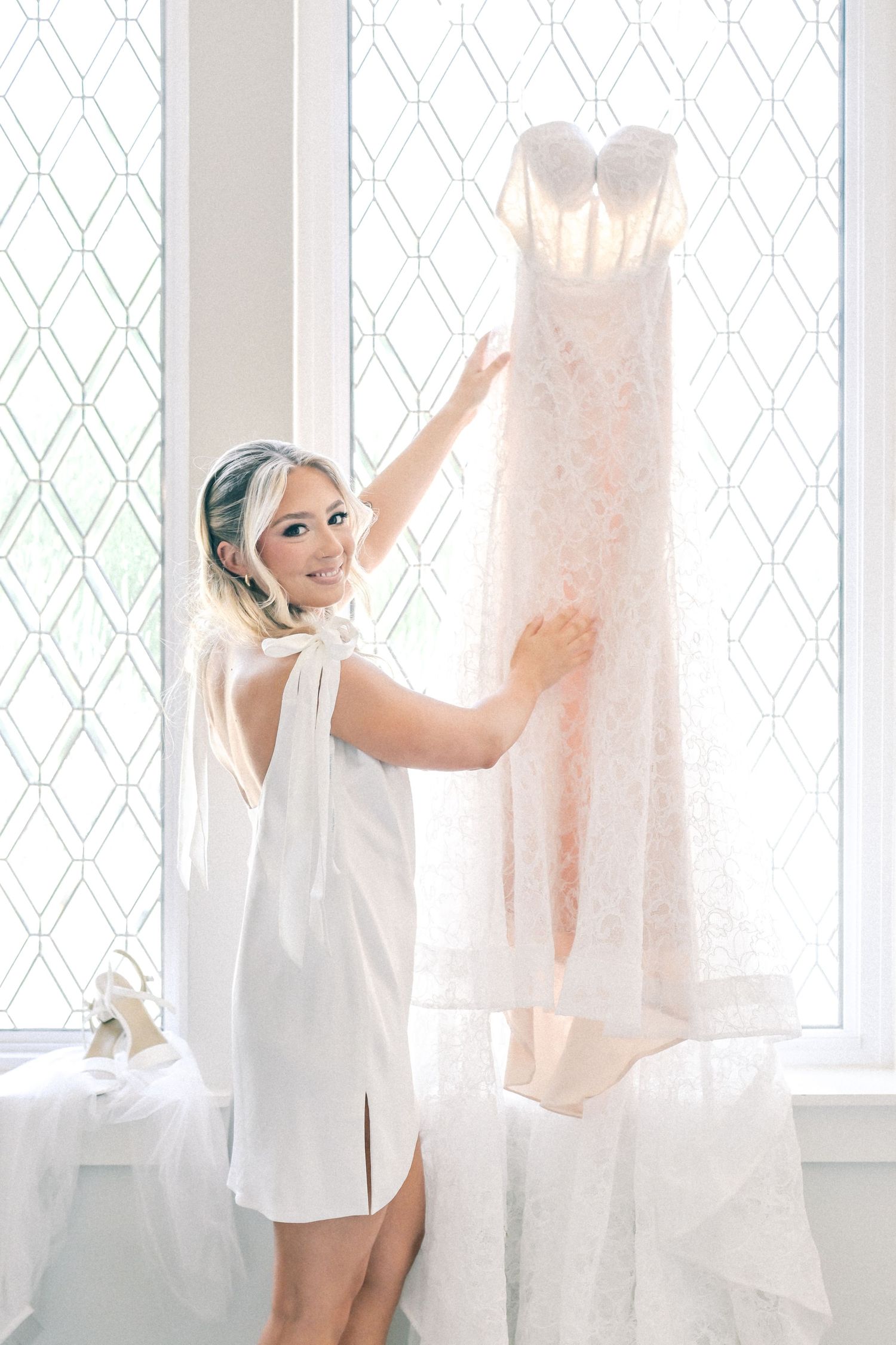 Blonde bride in white robe admiring her lace wedding dress hanging by a bright latticed window.