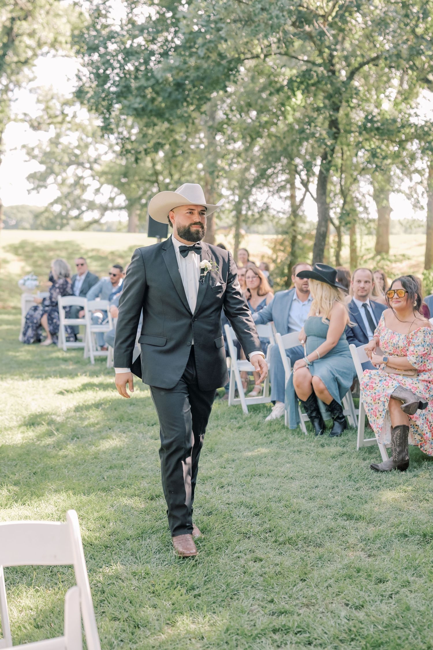 Groom in dark suit and cowboy hat walking down outdoor wedding aisle with seated guests.