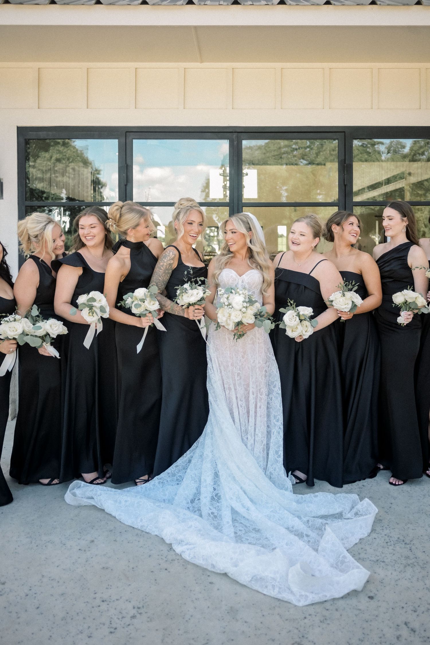 Bride in lace gown with bridesmaids in black dresses holding white bouquets outside wedding venue.