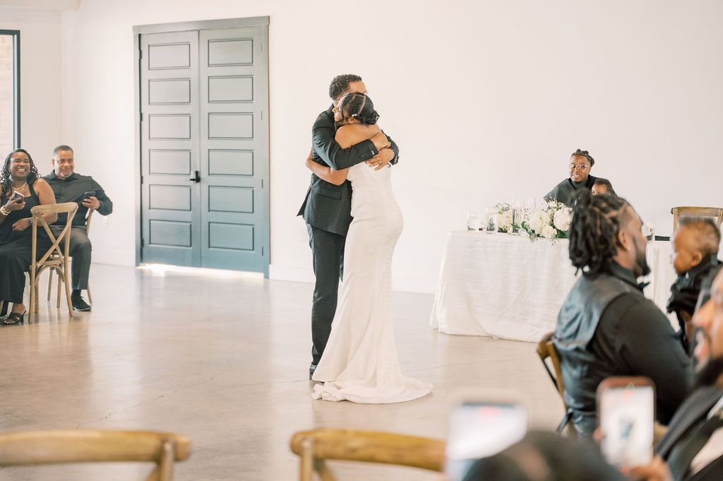 Newlyweds share their first dance as smiling guests look on in a modern white wedding reception space.