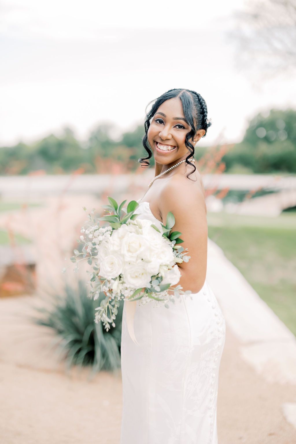 Smiling bride in strapless white gown holding elegant white rose bouquet with greenery outdoors.