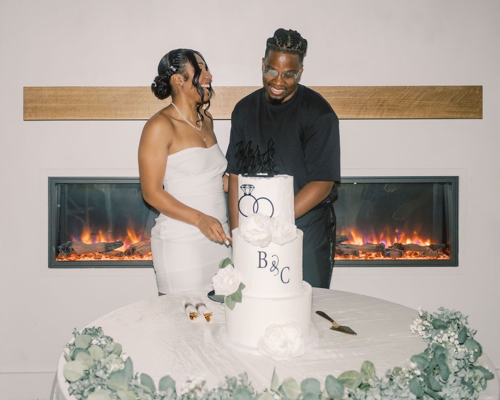 Couple cutting a white wedding cake with B&C monogram in front of a modern fireplace.