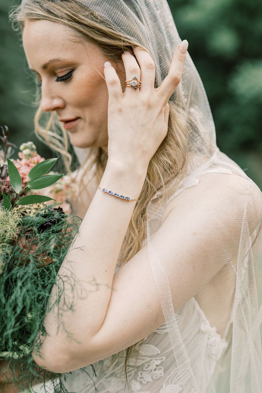 Bride in lace wedding dress and veil holding bouquet, showcasing diamond ring and bracelet.