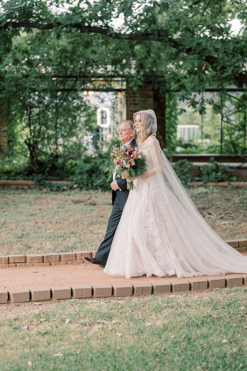 Bride in lace gown and veil walks with escort through garden arbor holding red floral bouquet.