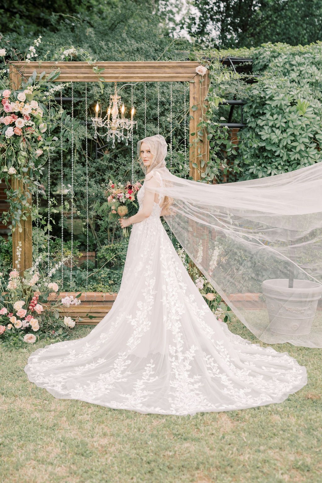 Bride in lace ballgown with flowing veil stands before floral arch with chandelier in garden.