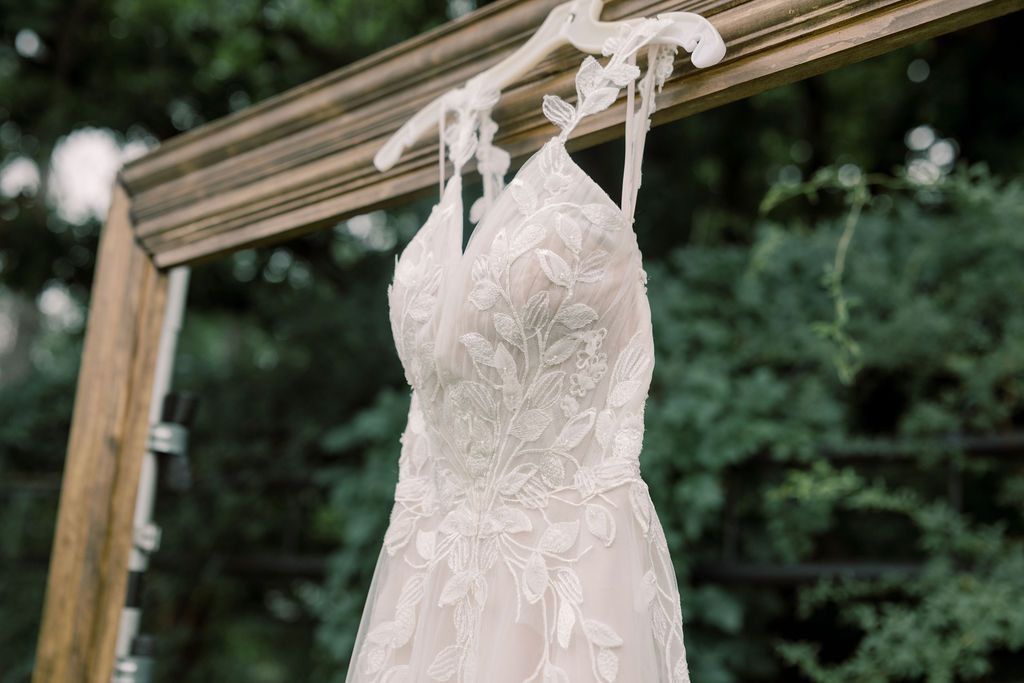A delicate lace wedding dress hanging on a white hanger against a rustic wooden frame outdoors.