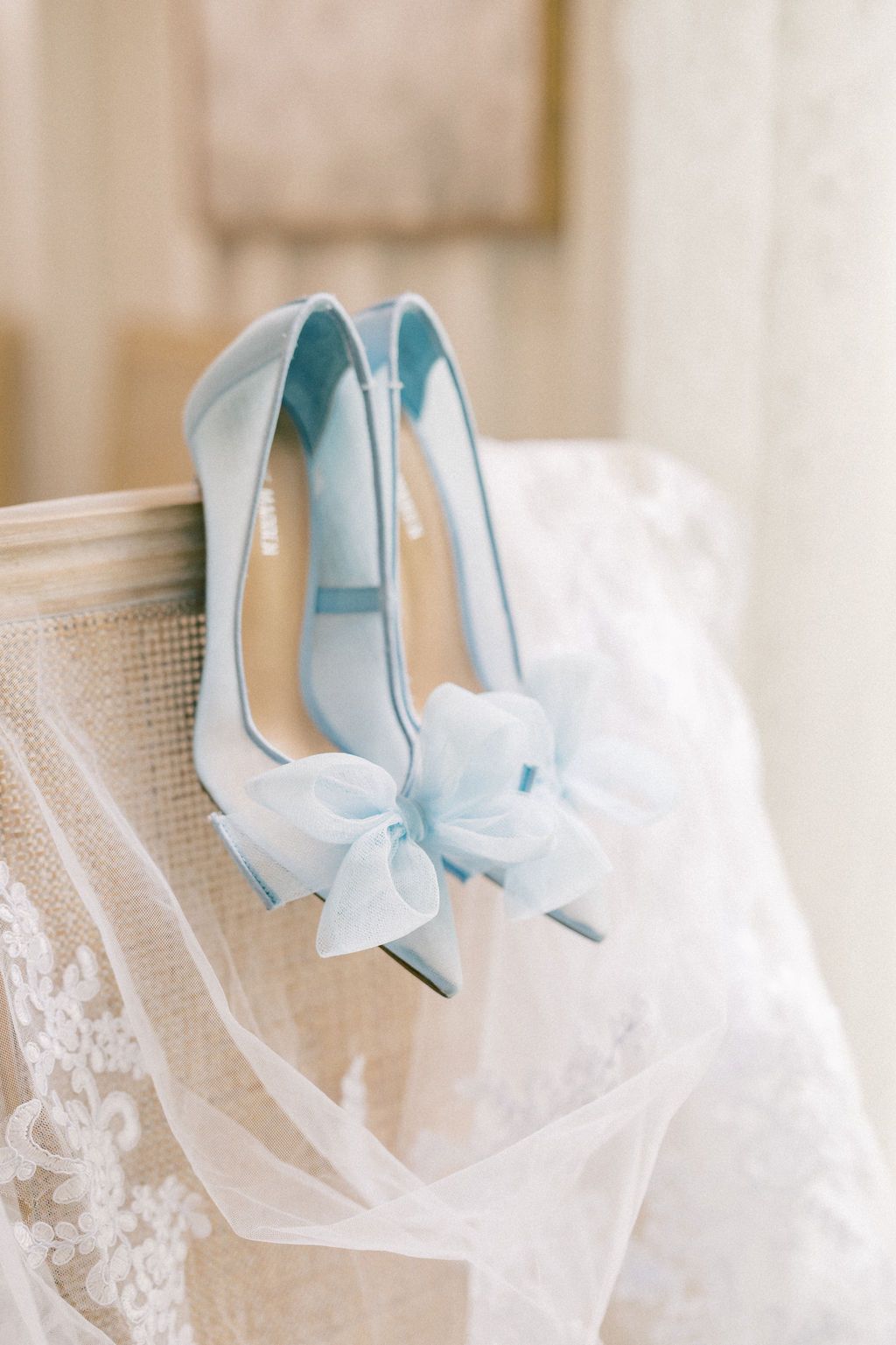 Delicate light blue bridal heels with bow detail resting on a lace wedding veil on a vintage chair.
