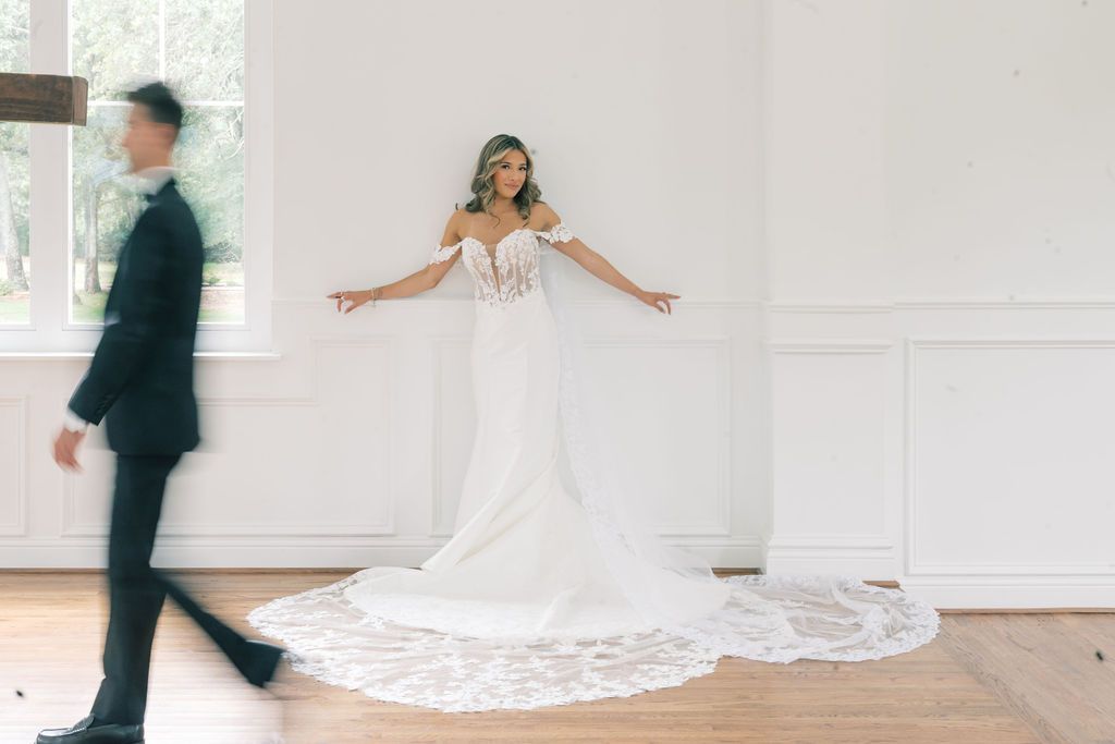 Bride in strapless lace wedding gown with long train spreads arms wide as groom walks toward her.